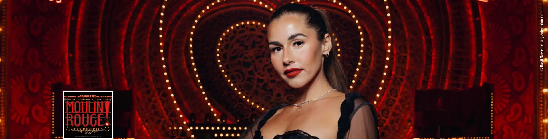 Frau / Sarah Engels mit langen dunklen Haaren trägt ein schwarzes, transparentes Oberteil mit Rüschen vor einem roten, leuchtenden Herzmuster und einem 'Moulin Rouge!' Schild.