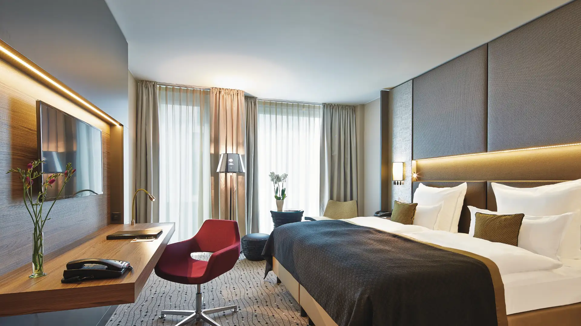 Steigenberger Hotels im Kachelformat: Modernes Hotelzimmer im Steigenberger Hotel am Kanzleramt mit großem Doppelbett, brauner Tagesdecke, rotem Stuhl und großem Fenster mit Vorhängen.