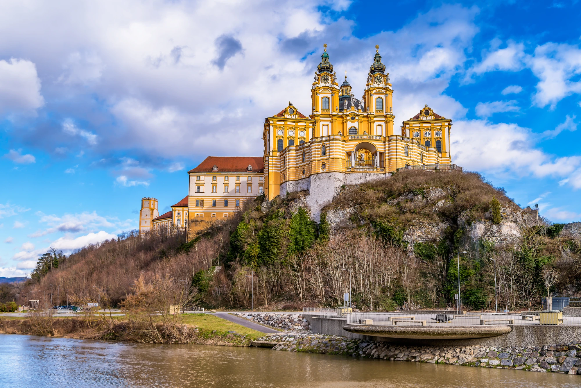 Gelb-weiße barocke Melker Stiftskirche auf einem bewaldeten Hügel mit Fluss und Brücke im Vordergrund unter blauem Himmel mit Wolken