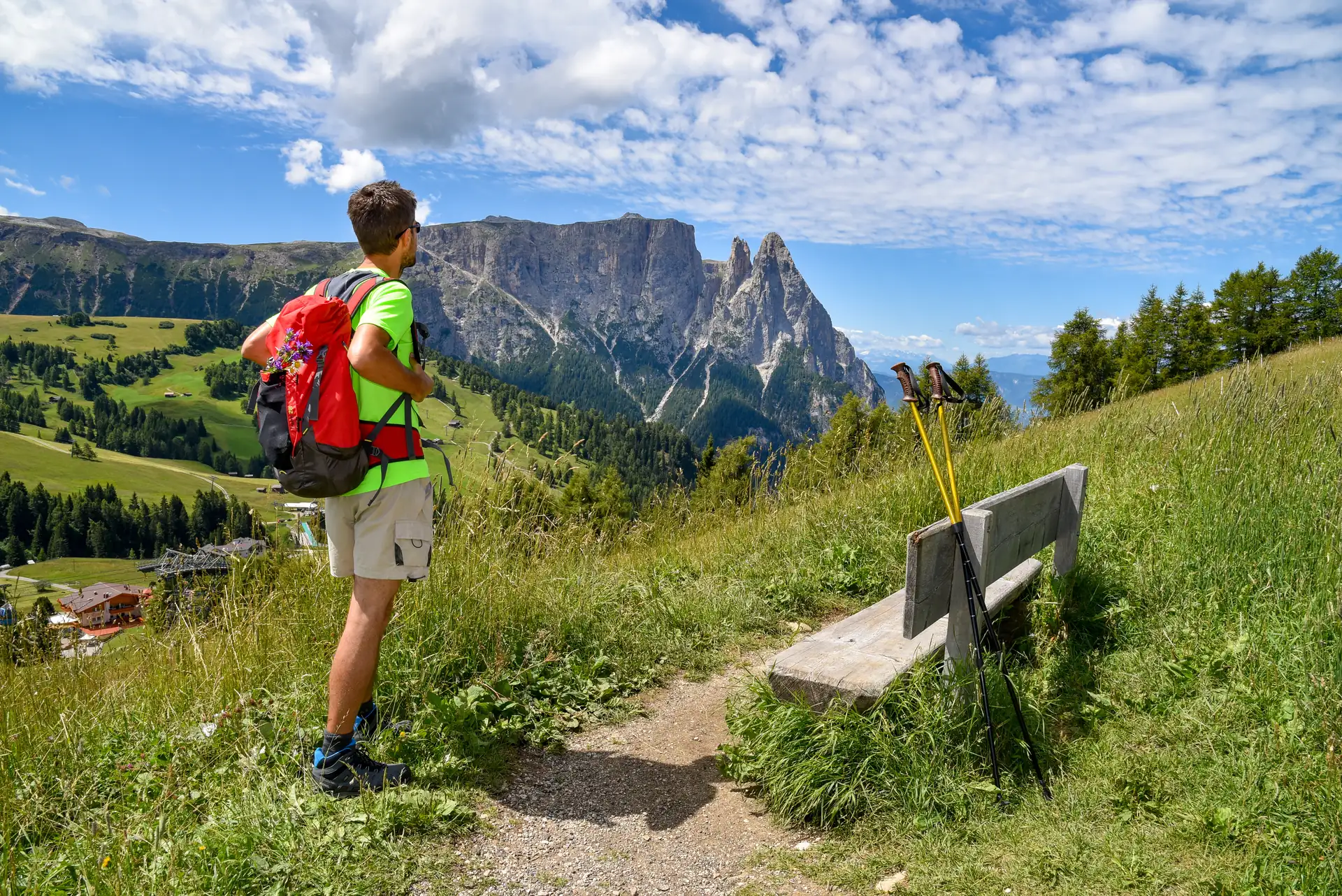 Wanderer mit rotem Rucksack steht auf einem Pfad und blickt auf die Bergspitzen der Seiser Alm, neben einer Bank mit Wanderstöcken.