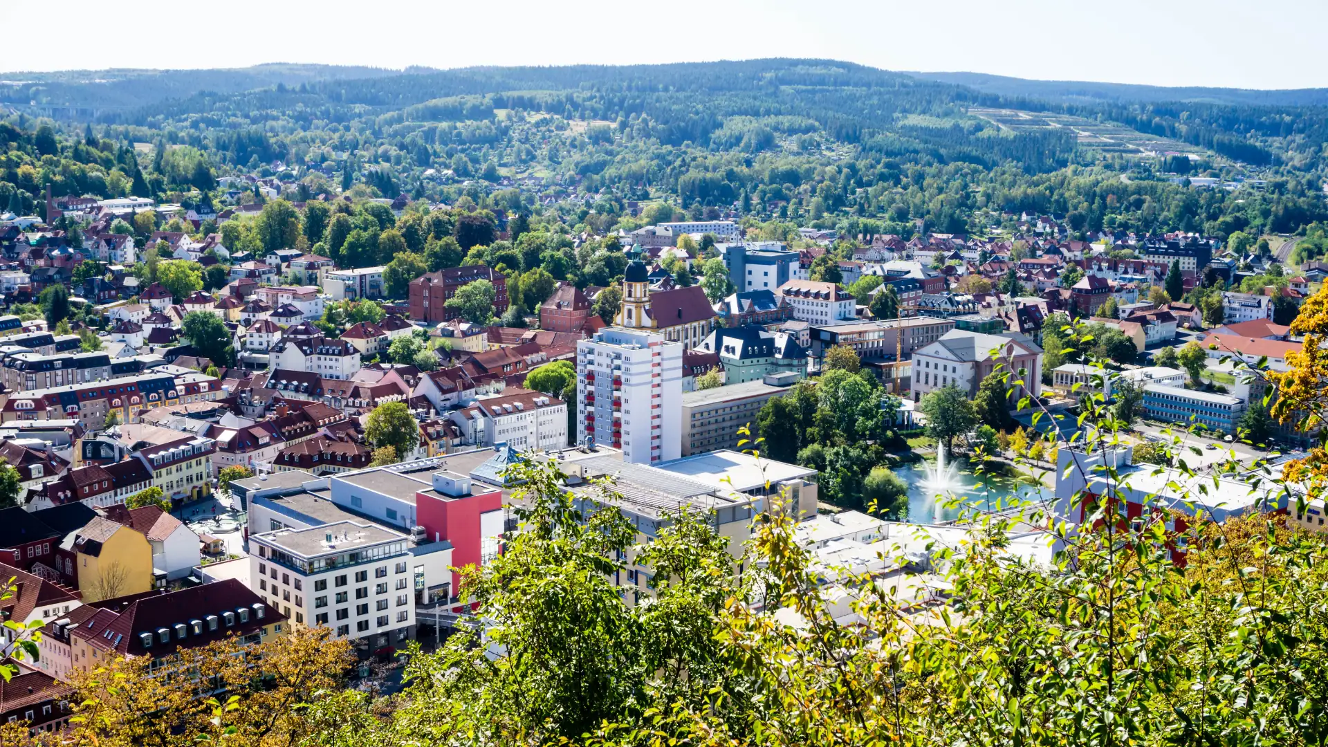 Luftaufnahme der Stadt Suhl mit vielen Gebäuden, einem Turm mit Uhr und einem See mit Springbrunnen im Vordergrund