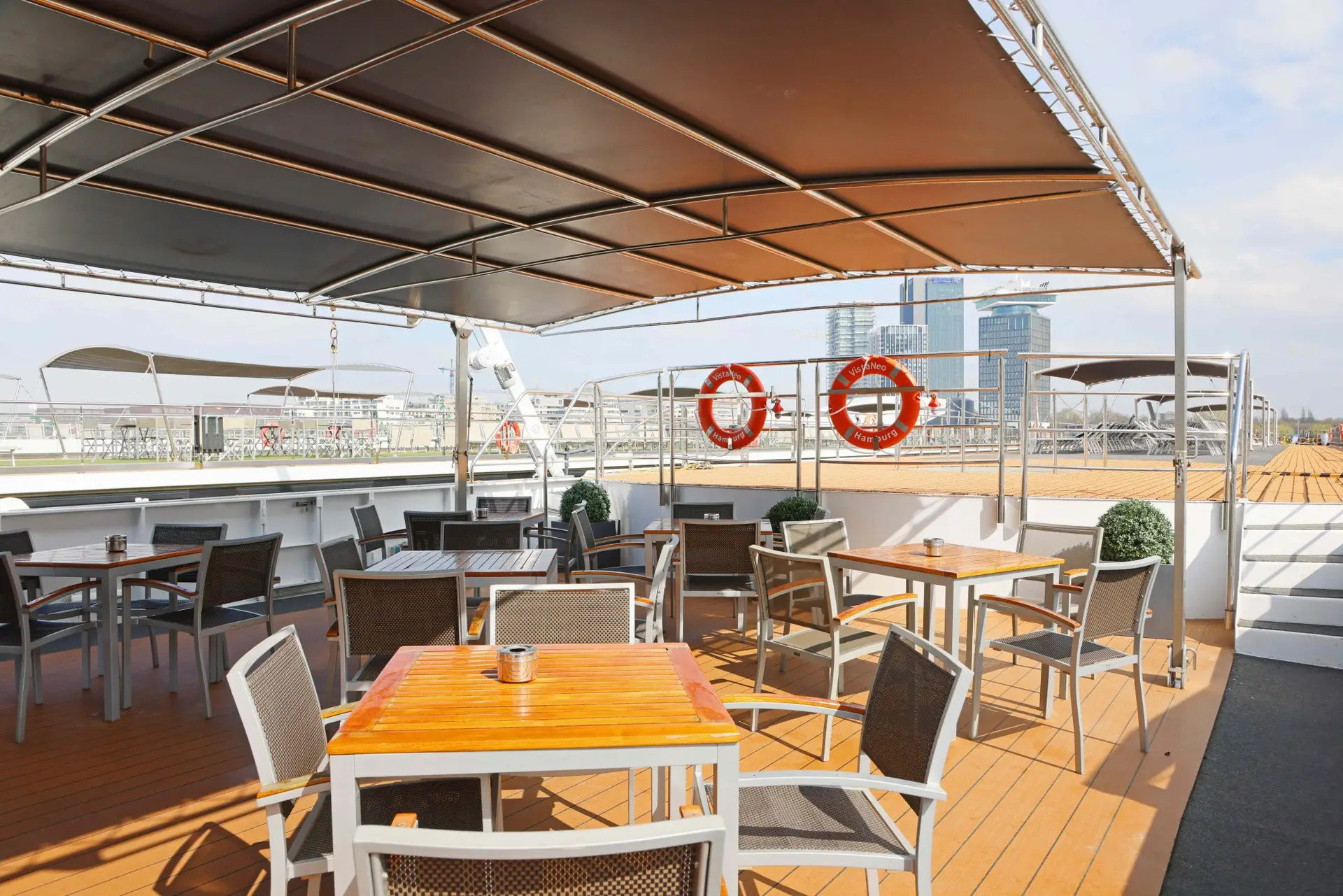 Überdachte Außenterrasse mit mehreren Tischen und Stühlen, zwei Rettungsringen am Geländer, Hochhäuser im Hintergrund