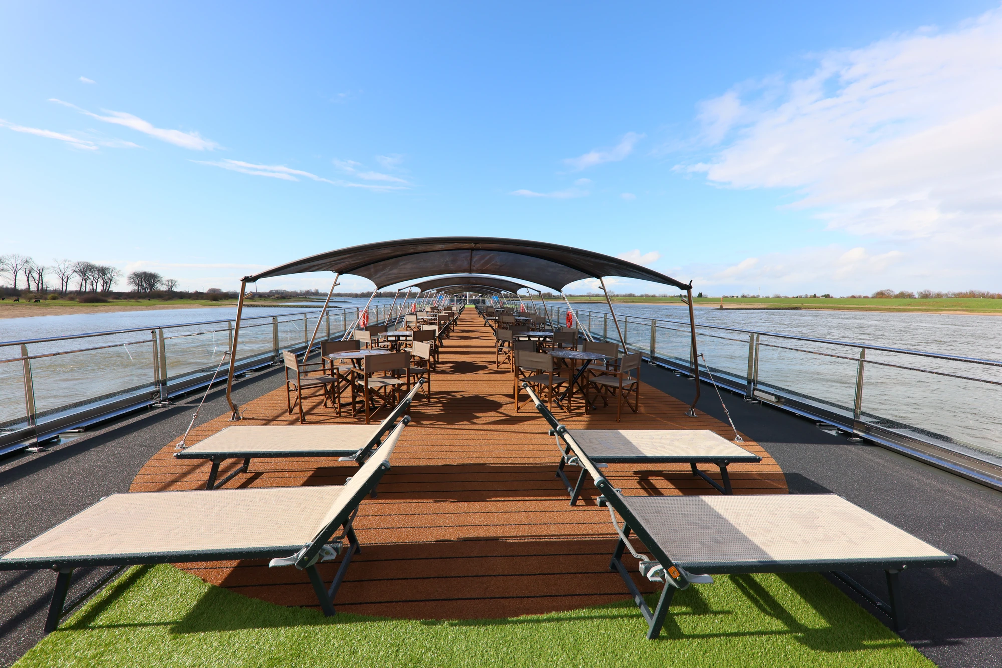 Sonnendeck eines Schiffes mit Liegestühlen, Tischen und Stühlen unter einem großen Sonnendach, Fluss und Ufer im Hintergrund