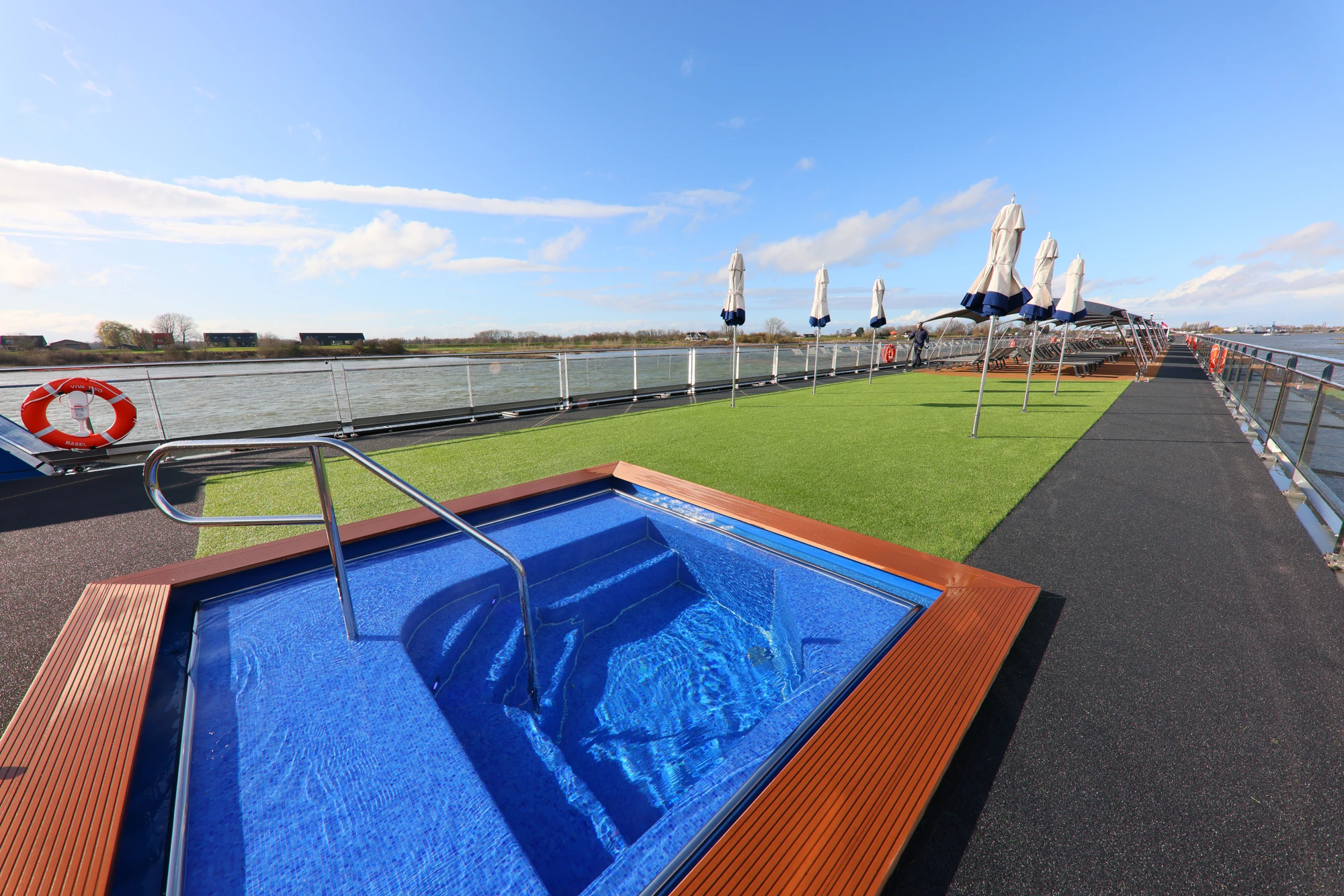 Blauer Whirlpool mit Holzrahmen auf einem Schiffssonnendeck mit grüner Kunstrasenfläche, weißen Sonnenschirmen und Wasser im Hintergrund.