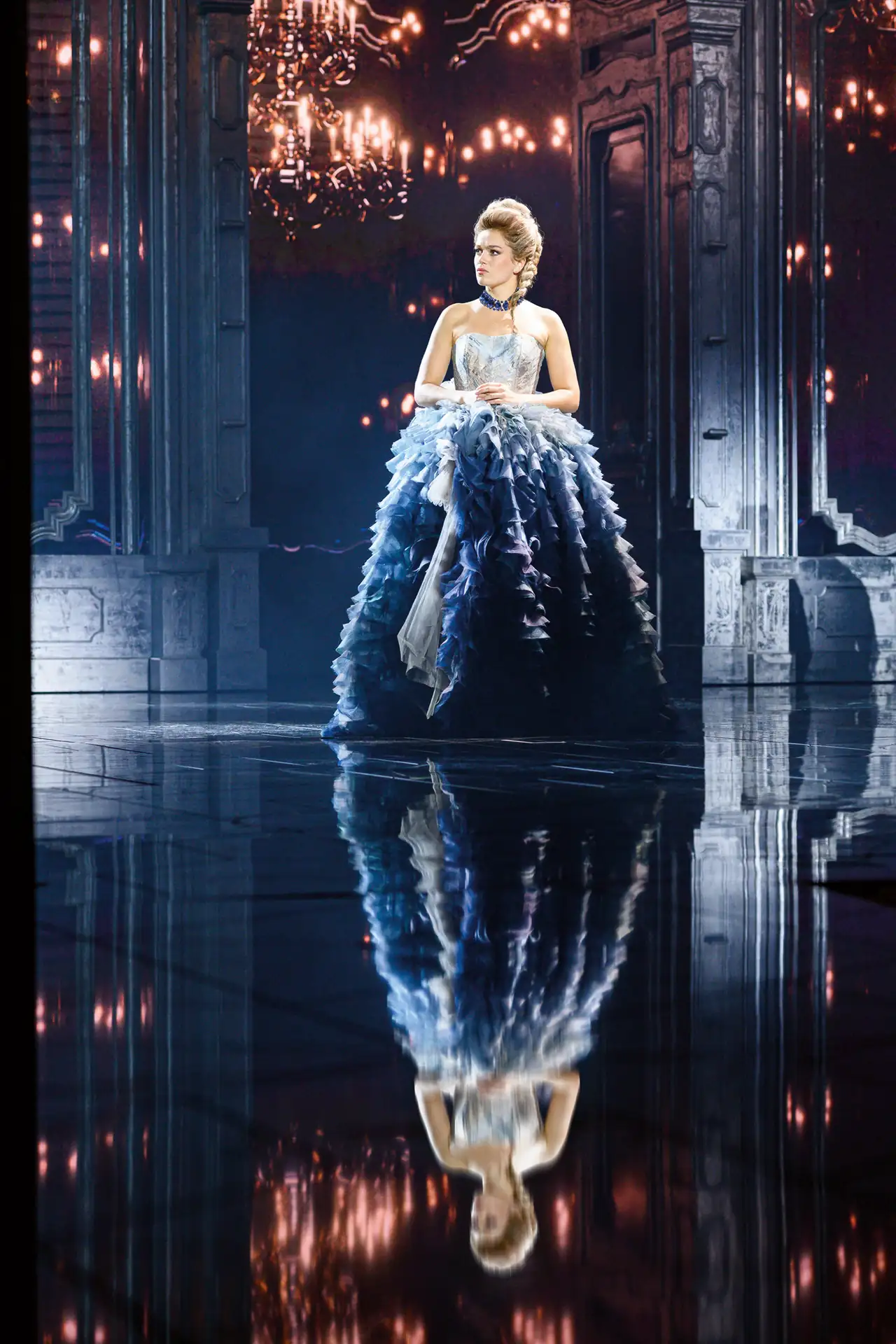 Maria Theresia Musical Szenenfoto: Frau in bodenlangem, voluminösem Kleid mit Rüschen, stehend vor prunkvollem, dunklem Hintergrund mit Kronleuchter, Spiegelung auf glänzendem Boden.