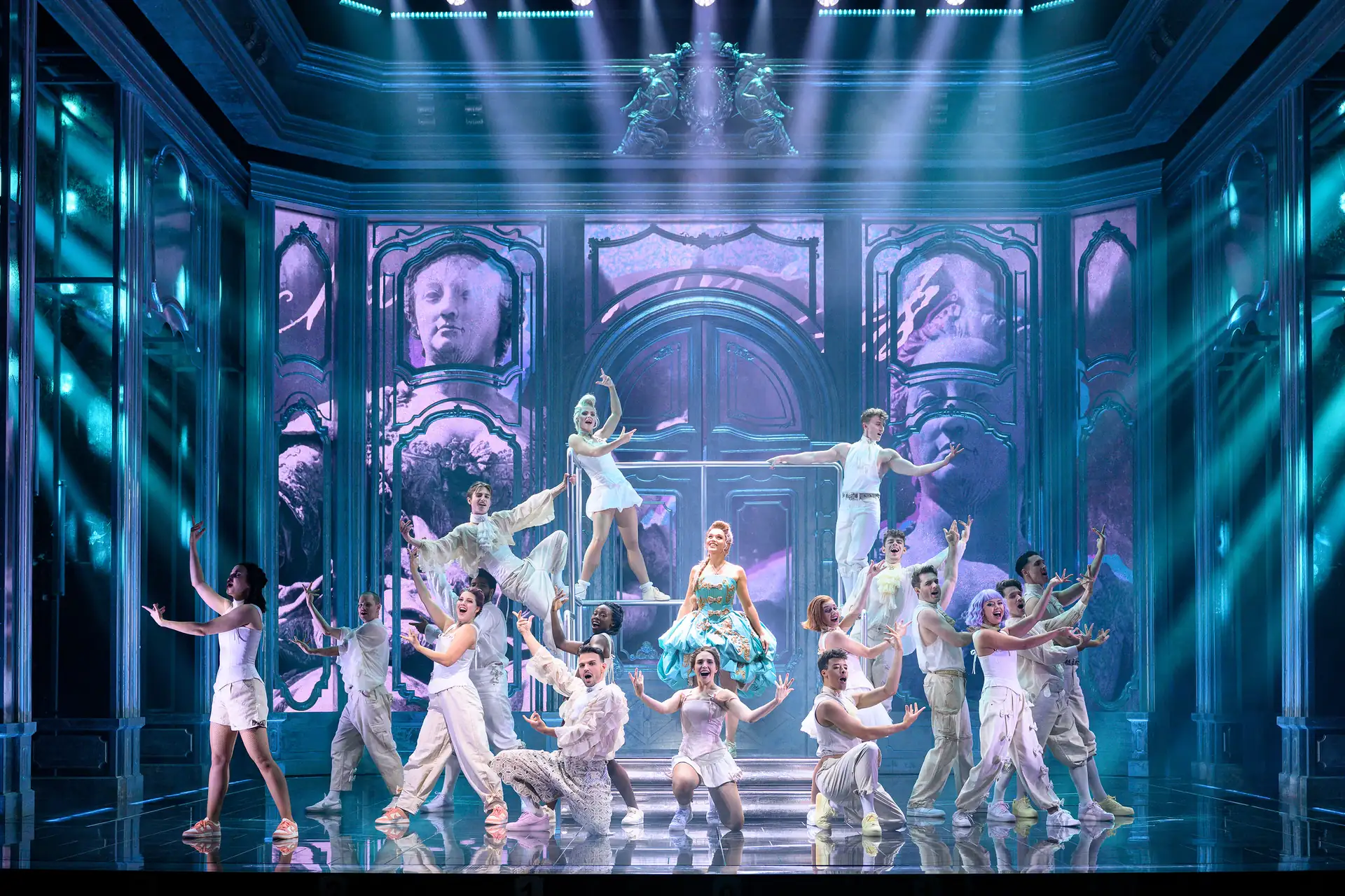 Maria Theresia Musical Szenenfoto: Bühnenszene mit Tänzern in weißen Kostümen, eine Tänzerin in blauem Kleid auf einer Treppe, barocke Kulisse mit großen Türen und Säulen.