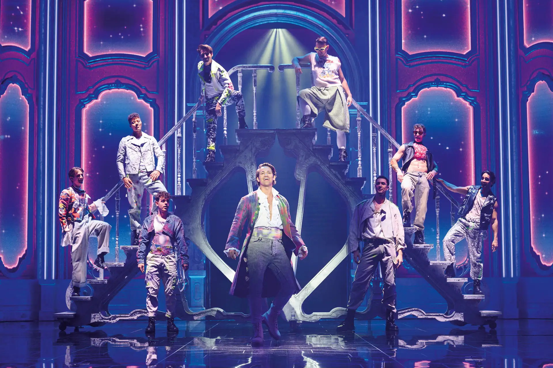 Maria Theresia Musical Szenenfoto: Acht Personen in bunter Bühnenkleidung stehen auf einer Treppe vor einer leuchtenden, barock inspirierten Kulisse mit blauen und pinken Lichteffekten.