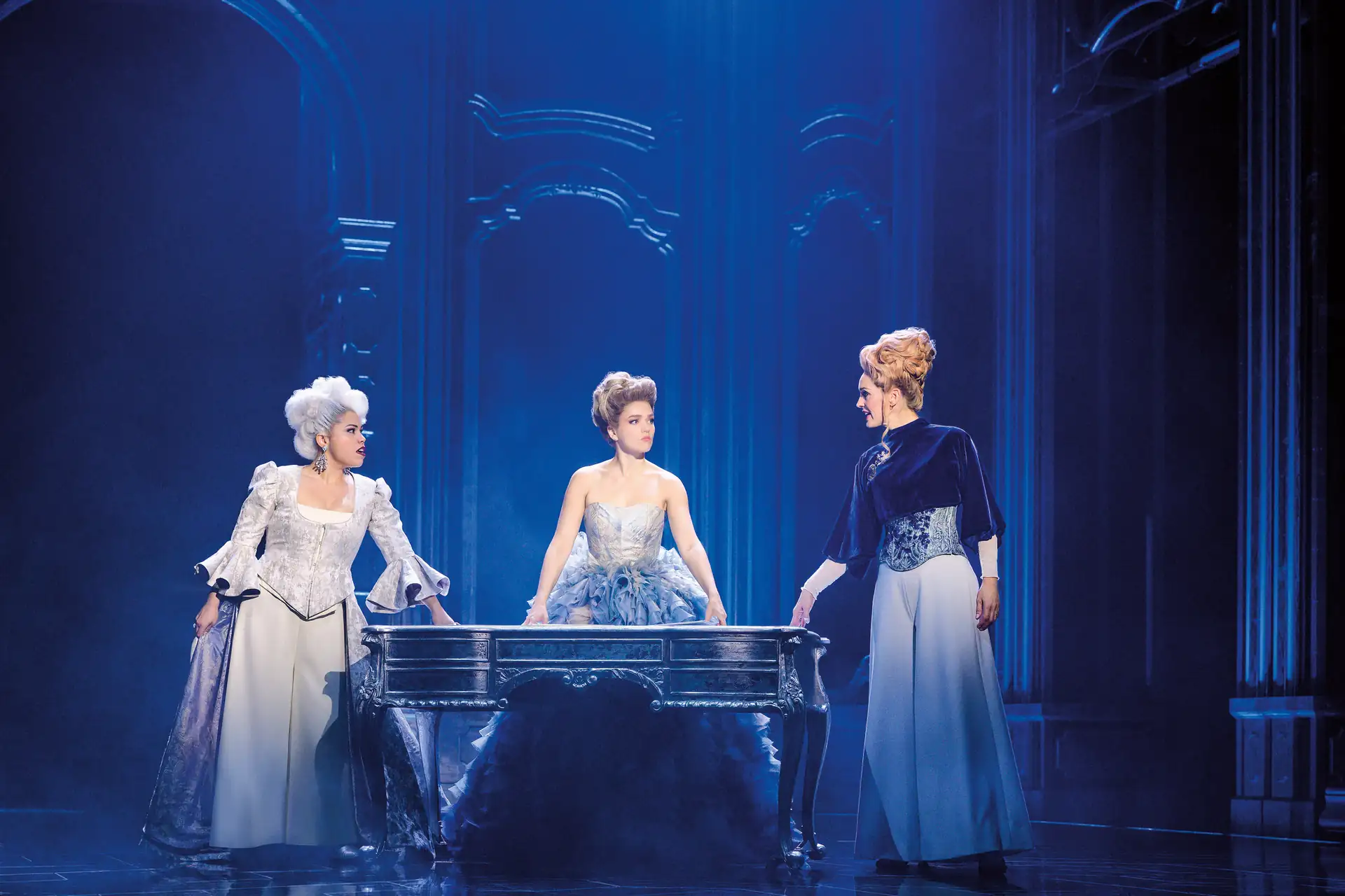 Maria Theresia Musical Szenenfoto: Drei Frauen in historischen Kostümen stehen um einen verzierten Tisch in einem prunkvollen, blau beleuchteten Raum.