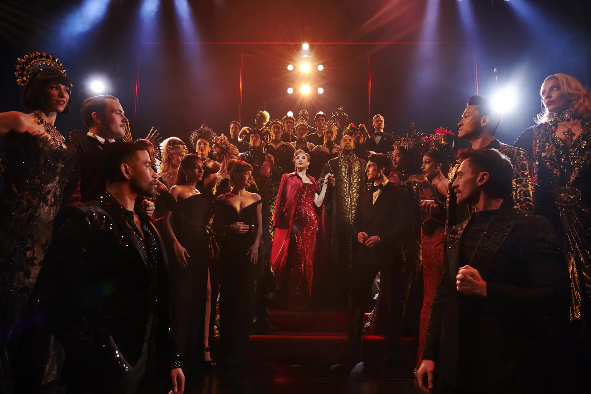 Ensemble des UK-Casts von »Der Teufel trägt Prada Das Musical« in eleganter Bühnenkleidung vor dramatischer Beleuchtung auf einer Treppe versammelt