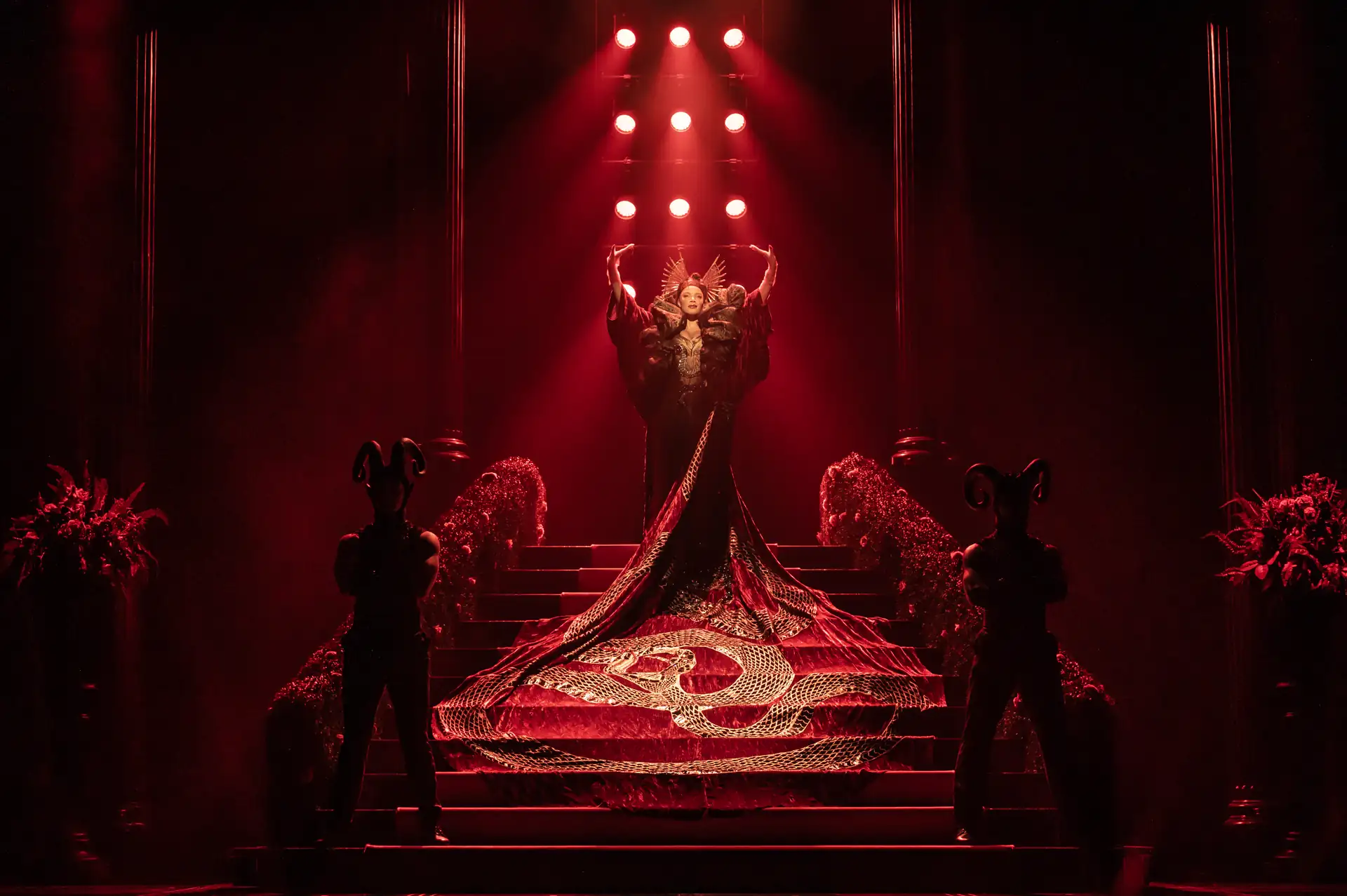 UK Cast vom Musical Der Teufel trägt Prada: Person in aufwändigem, langem Kleid mit Schlangenmuster steht auf einer Treppe vor roter Bühnenbeleuchtung, flankiert von zwei Personen mit Hörnern auf dem Kopf.