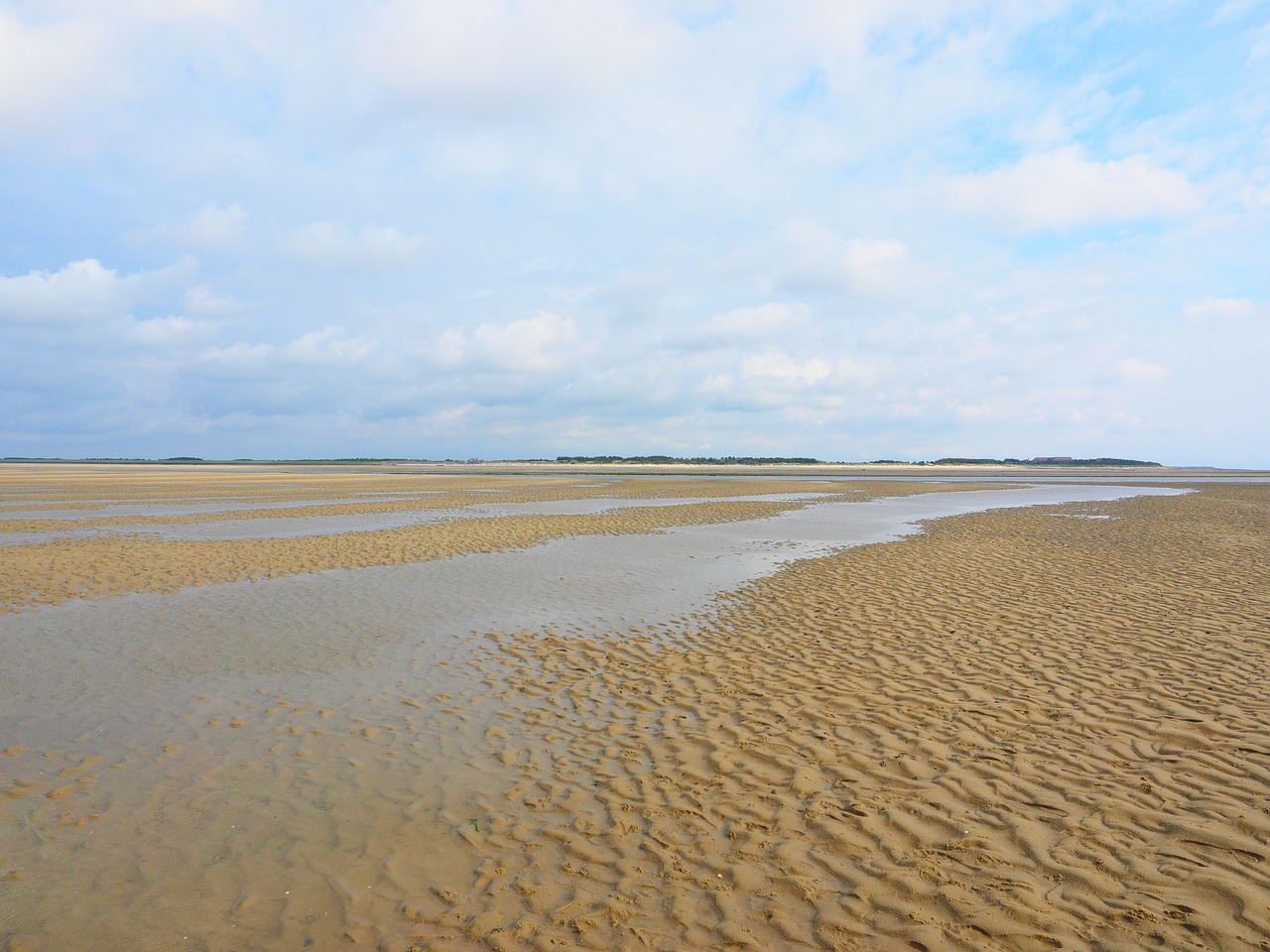 Weite Sicht auf einen ruhigen Sandstrand bei Ebbe, mit sanften Wellen und weichen Wolken am blauen Himmel.