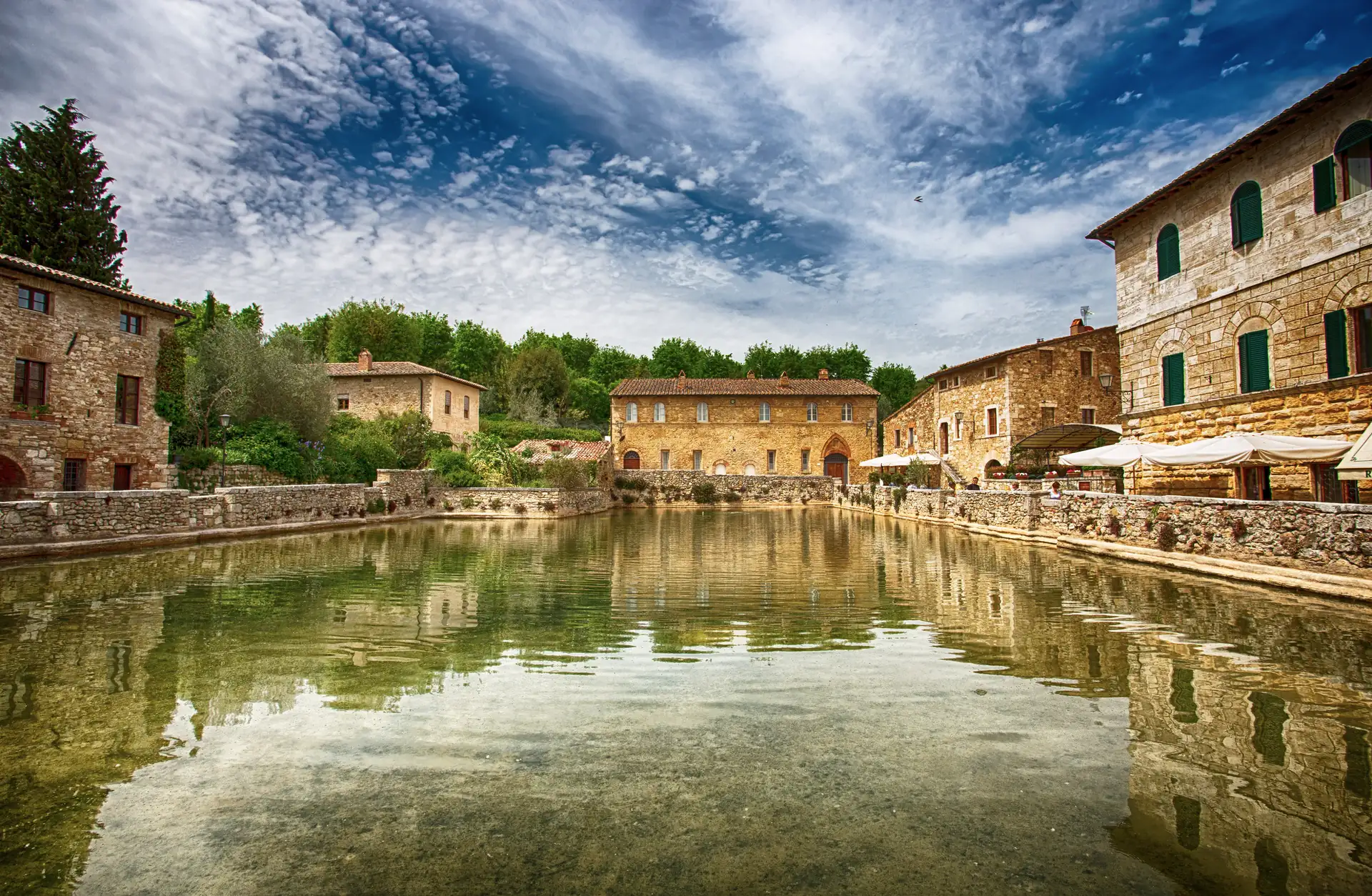 Bagno Vignoni, historisches Dorf in der Toskana mit Thermalbädern, Steinhäusern und grünem Bewuchs um einen großen, ruhigen Wasserteich unter bewölktem Himmel