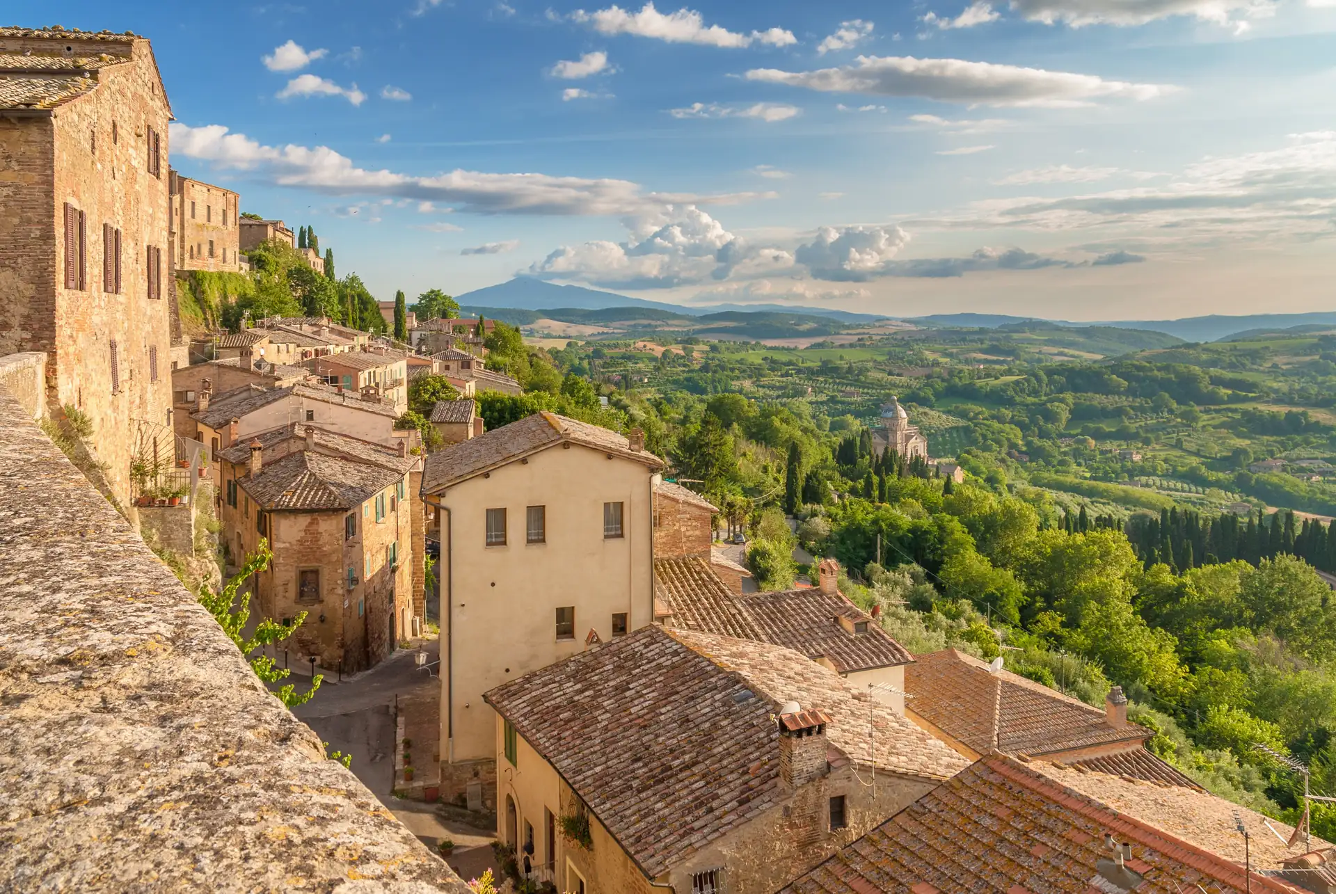 Blick von einer Stadtmauer auf Dächer des historischen Ortes Montepulciano in der Toskana mit Hügeln und bewaldeter Landschaft im Hintergrund