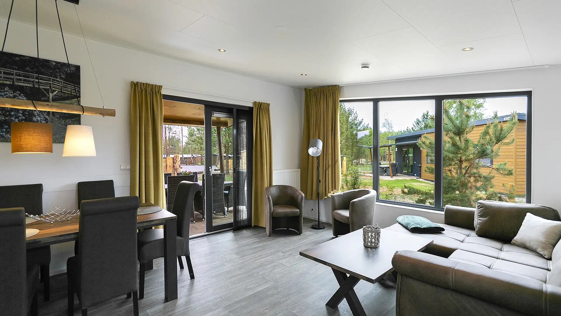 Innenansicht vom Sunrise Home: Helles Wohnzimmer mit grauem Sofa, Holztisch, Essbereich mit schwarzen Stühlen und großen Fenstern mit Blick auf Garten und Holzhaus.