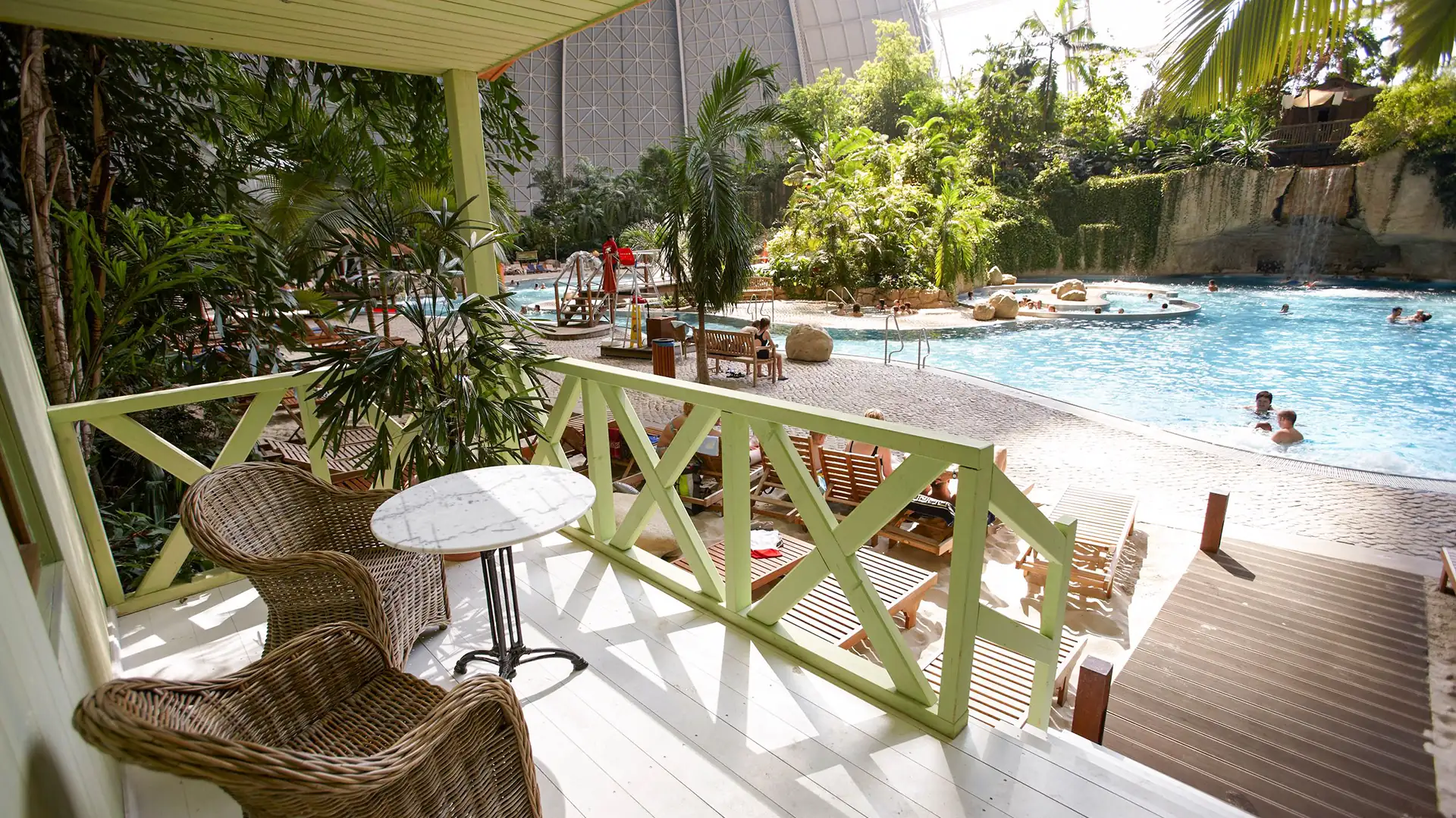 Tropical Islands: Blick von einer Terrasse mit zwei Korbsesseln und kleinem Tisch auf einen Pool mit Menschen und tropischer Vegetation.