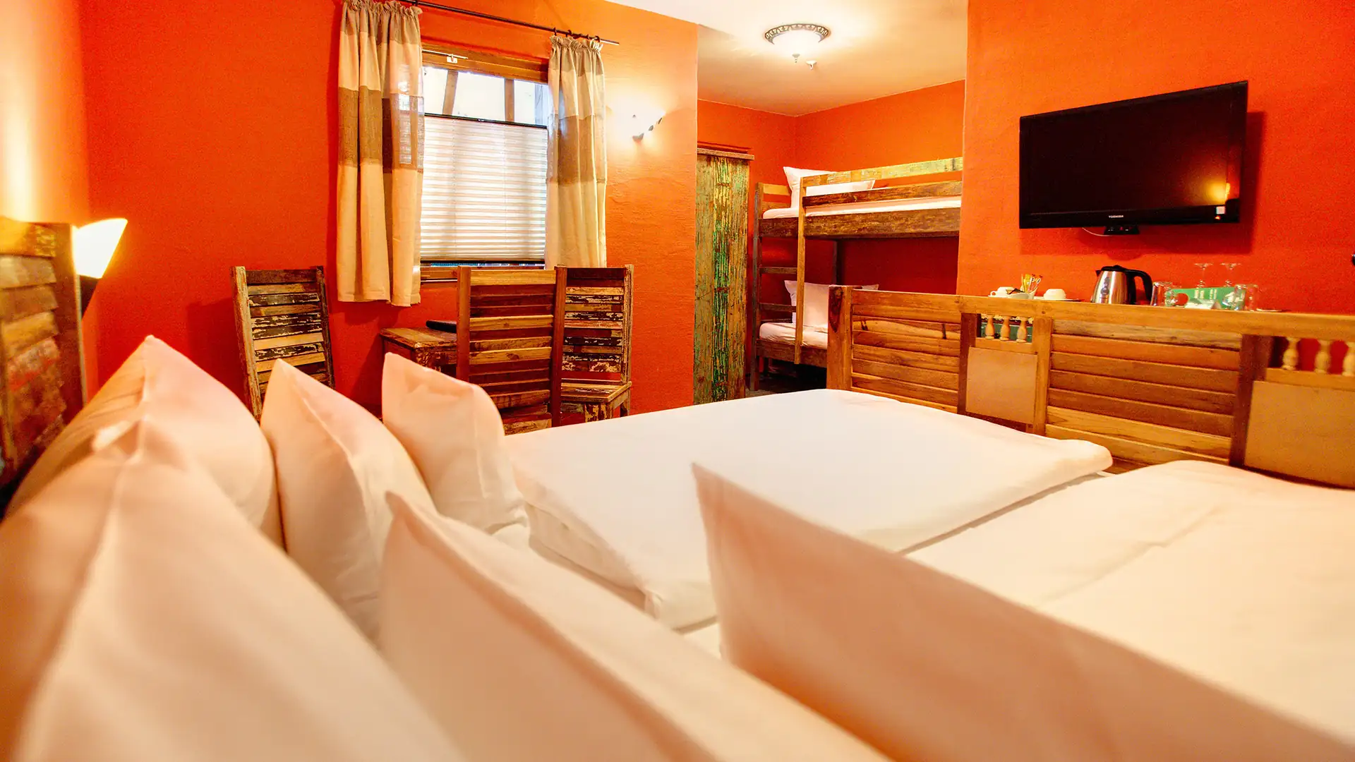 Hotelzimmer im Tropical Islands mit zwei Einzelbetten nebeneinander, Etagenbett, orange gestrichene Wände, Wandfernseher und Fenster mit Vorhängen