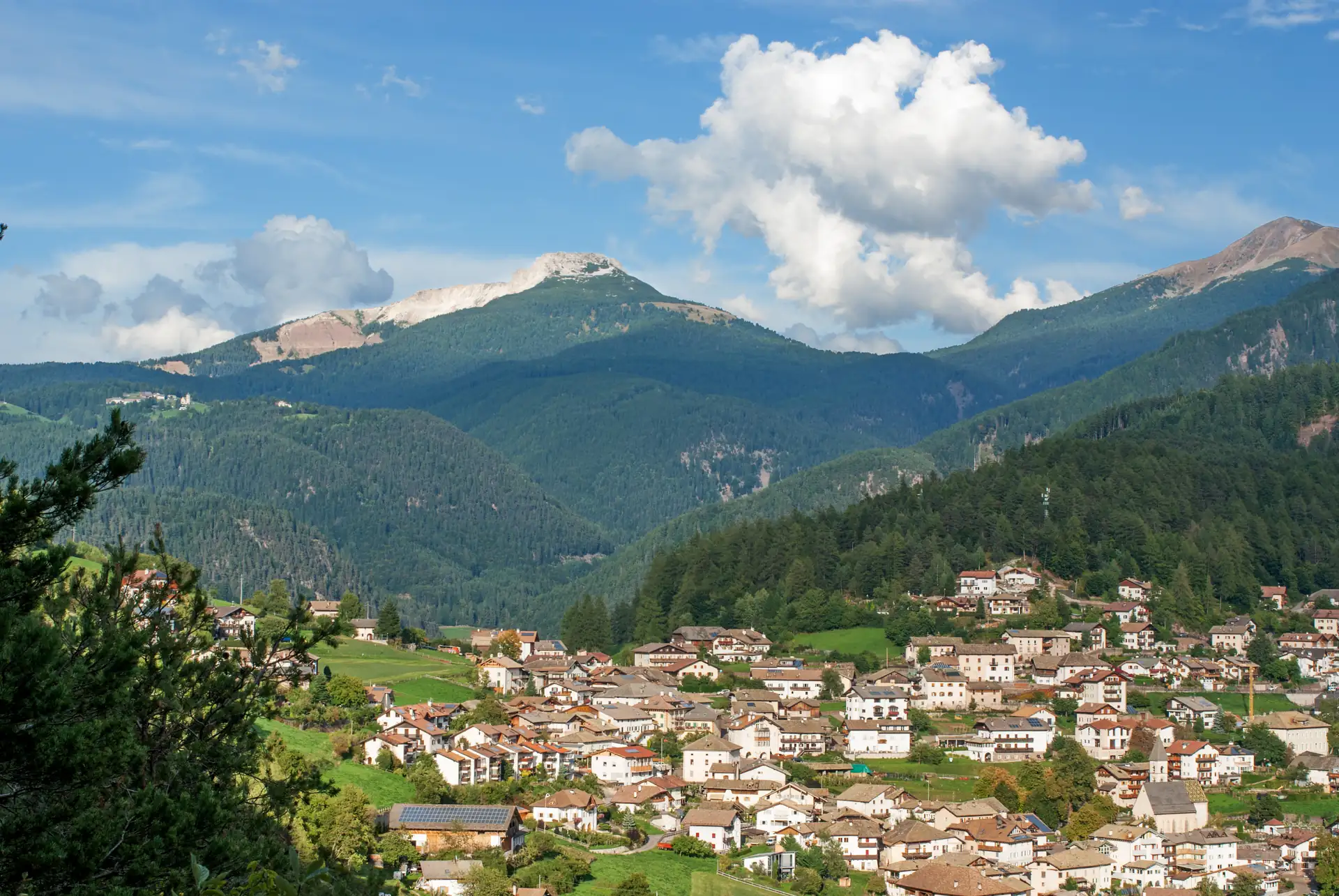 Bergdorf Truden mit zahlreichen Häusern vor bewaldeten Bergen und wolkigem Himmel in den Dolomiten.