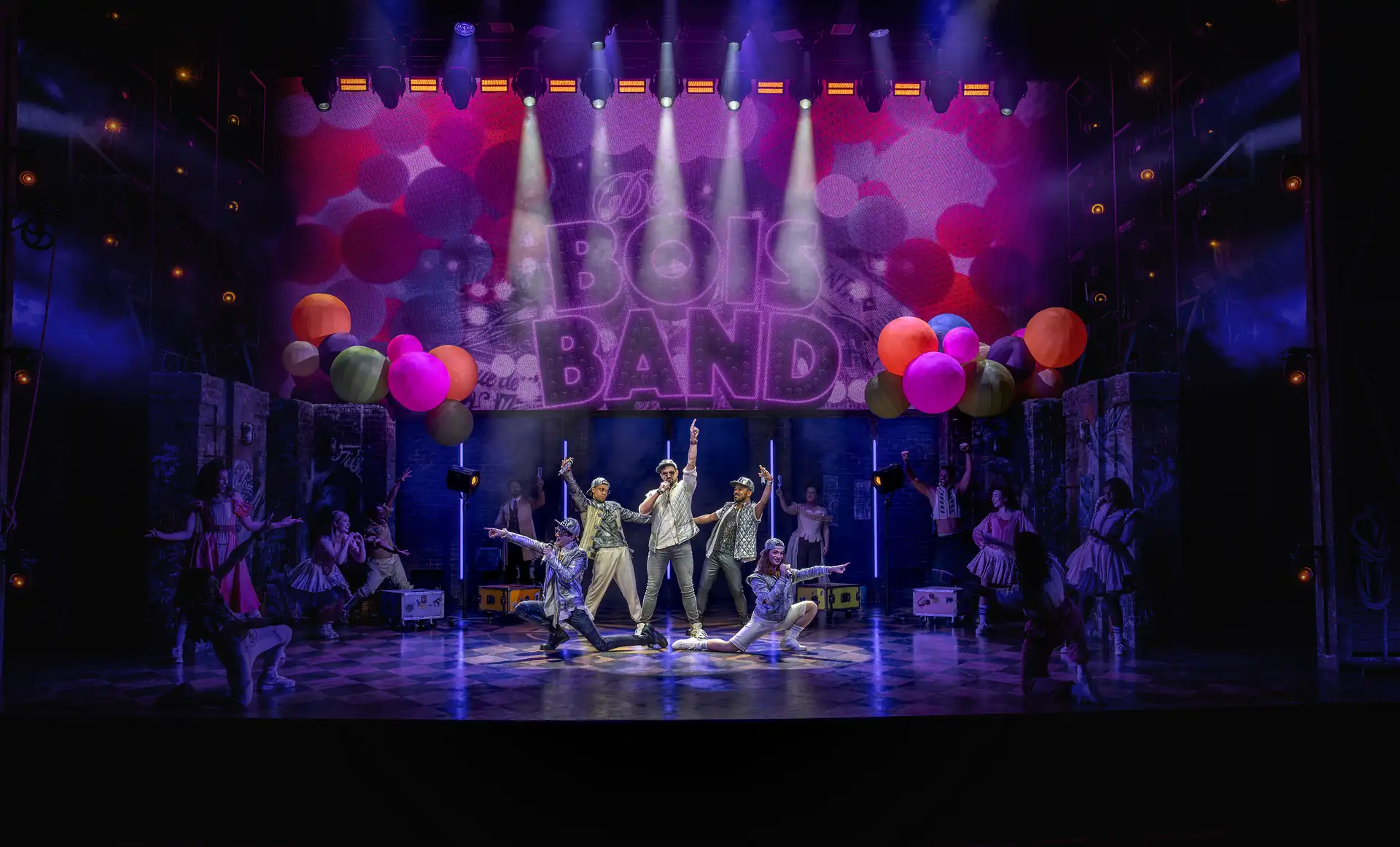 Bühnenbild des Julia-Musicals in Stuttgart mit einer Gruppe von sieben Darstellern in glänzenden Kostümen vor einer beleuchteten Wand mit dem Schriftzug 'De BOIS BAND' und bunten Luftballons.