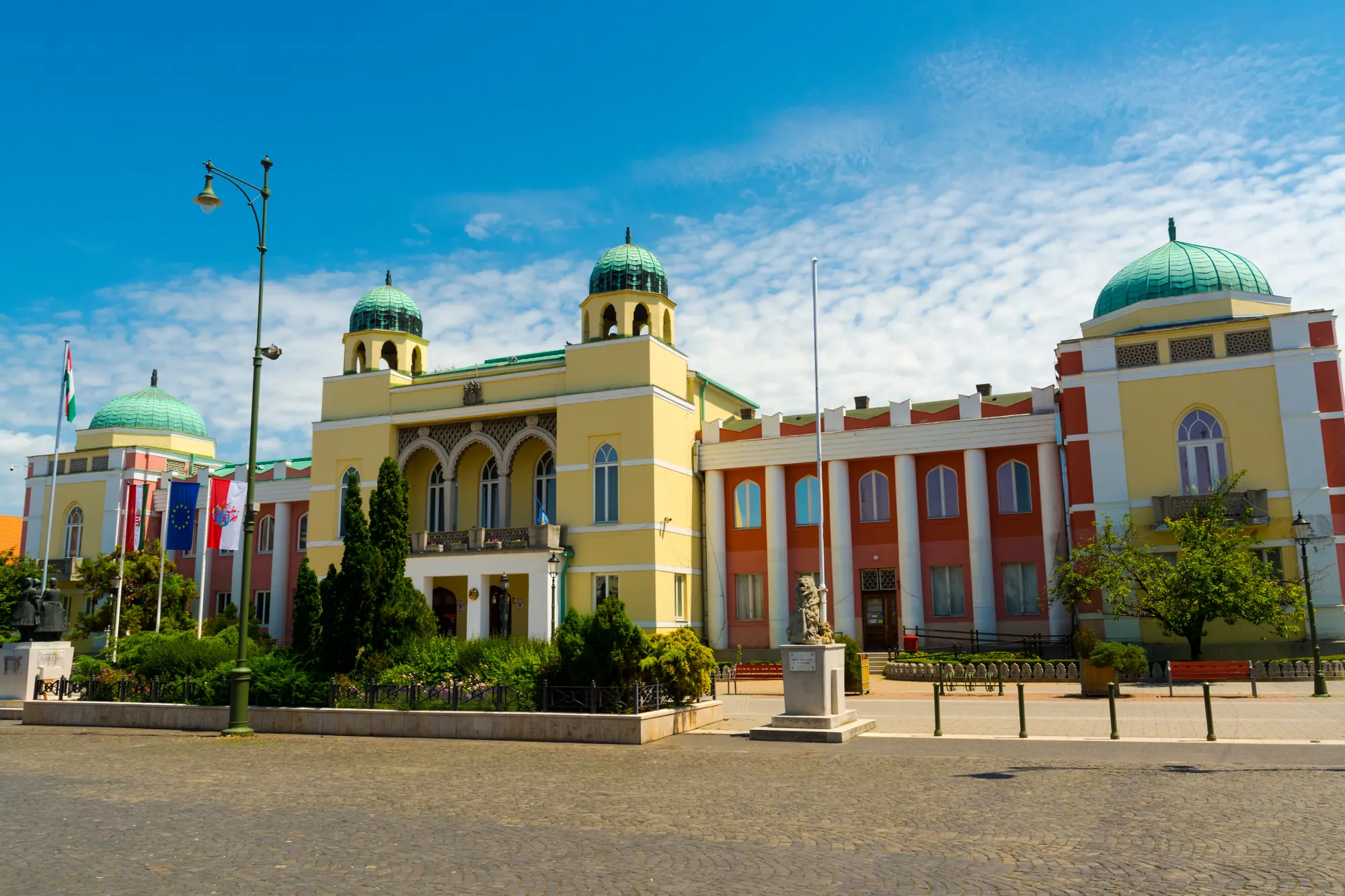 Rathaus in Mohács, Ungarn - farbenfrohes Gebäude mit grünen Kuppeln und architektonischen Details, umgeben von Gärten und Statuen unter einem strahlend blauen Himmel.