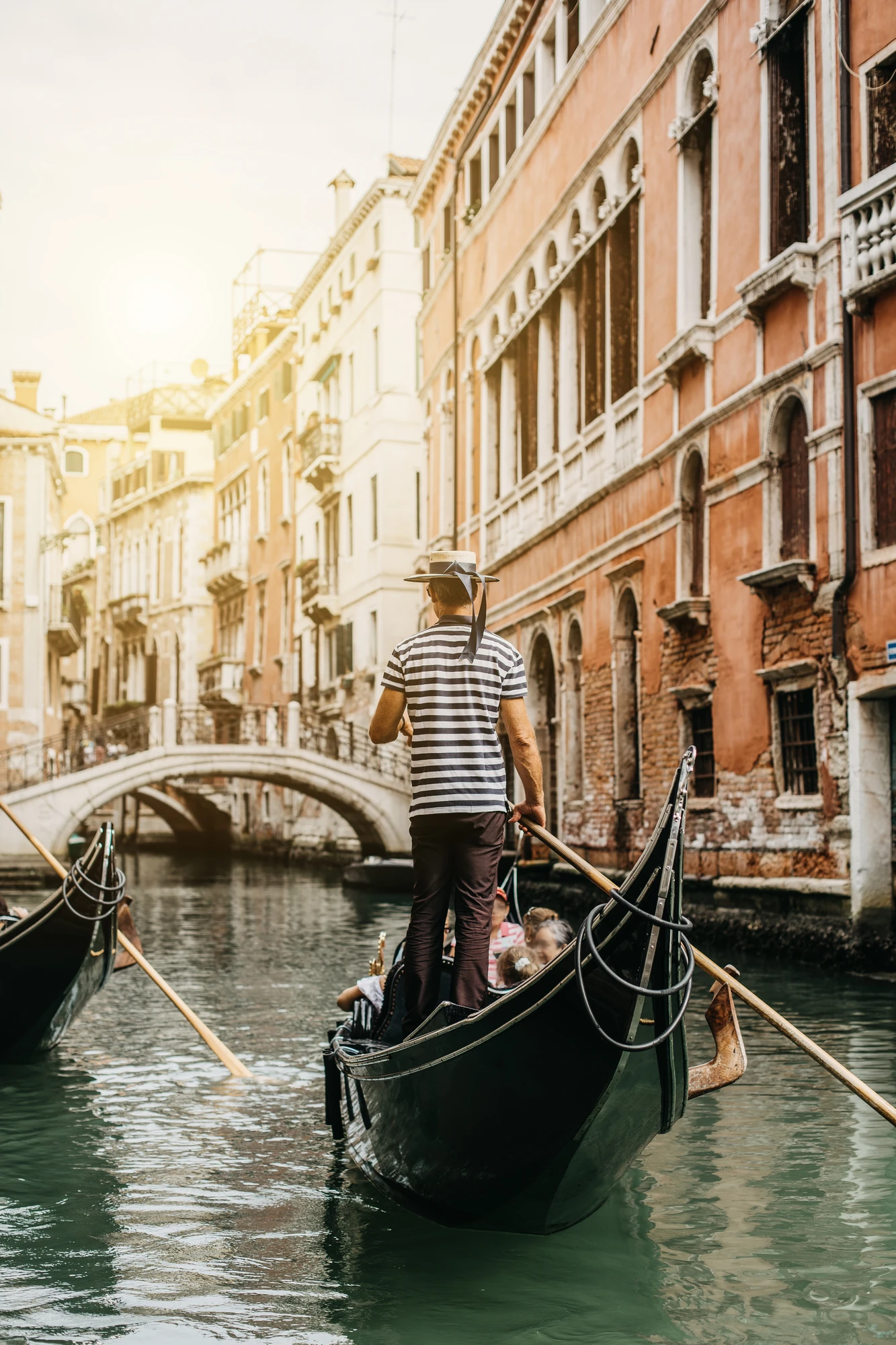 Gondoliere von hinten in den malerischen Wasserstraßen Venedigs. Die Szenerie zeigt die traditionellen Gondeln, während historische Gebäude entlang der Kanäle im Vordergrund sichtbar sind. Diese Aufnahme fängt das romantische Flair der Stadt ein und vermittelt ein Gefühl von Tradition und Eleganz.