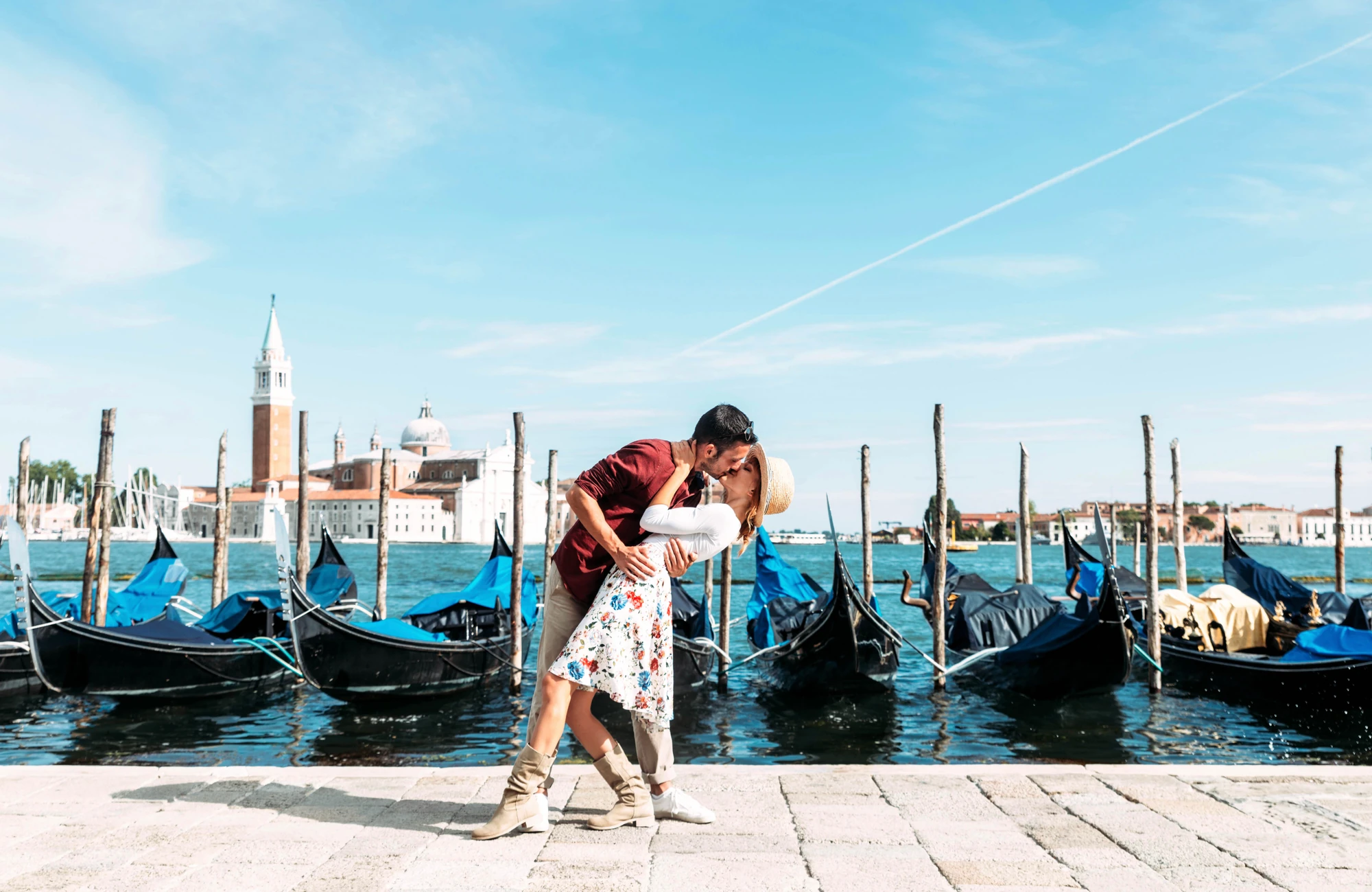 Ein Paar küsst sich romantisch vor den Gondeln an der Hafenpromenade in Venedig, umgeben von der charmanten Kulisse der Stadt. Das milde Wetter und die malerischen Wasserwege tragen zur idyllischen Atmosphäre bei und fangen den zauberhaften Moment perfekt ein.