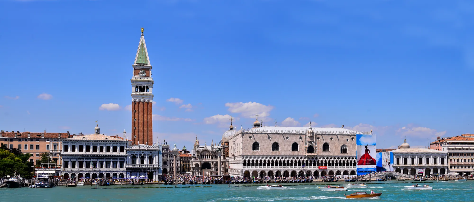 Panoramaansicht auf Venedig mit dem Fokus auf der südlichen Seite des Dogenpalastes, erhellt von klarem Tageslicht. Die beeindruckende Architektur des Palastes hebt sich vor dem strahlend blauen Himmel ab, während die umliegenden Wasserstraßen und historischen Gebäude die charakteristische Schönheit der Stadt unterstreichen.