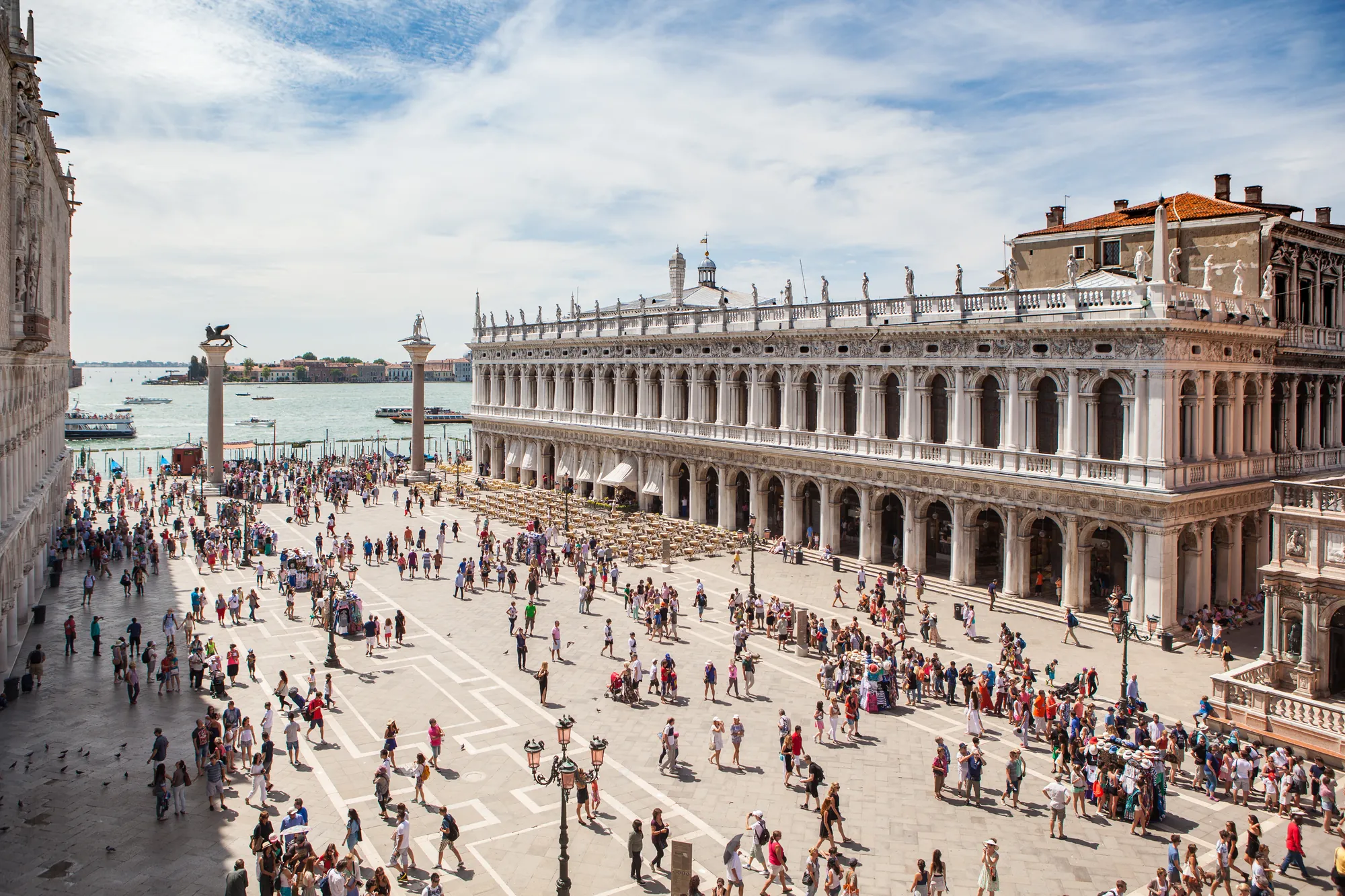 Seitlicher Blick auf die Marciana Bibliothek gegenüber dem Dogenpalast bei strahlendem Sonnenschein. Auf dem Marciana Platz tummeln sich viele Touristen, die Fotos machen, die beeindruckende Architektur bestaunen und gemütlich umherlaufen.