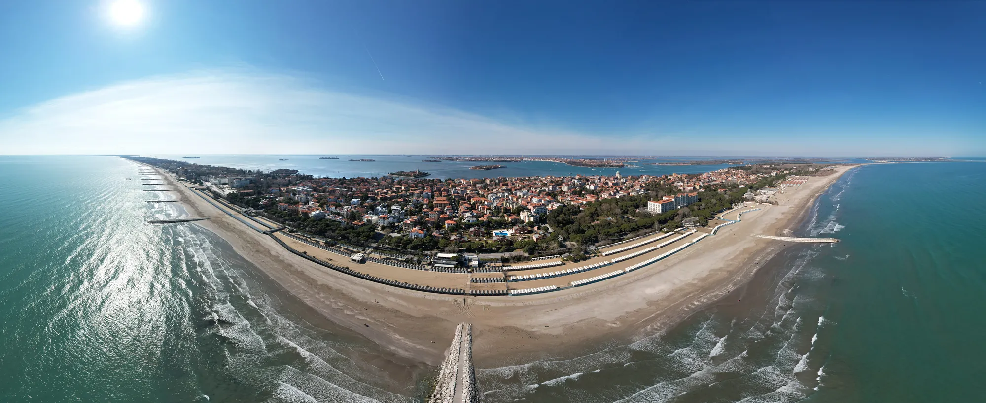 Panorama der Insel Lido di Venezia bei sonnigem und klarem Wetter, zeigt die gesamte Lagune in ihrem vollen Glanz. Umgeben von schöner Natur, erstreckt sich die Insel mit ihrem feinen Sandstrand und dem ruhigen, glitzernden Wasser, das sanft in die Lagune fließt.