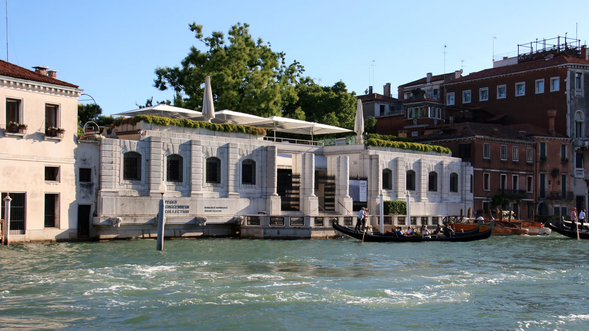 Seitliche Frontansicht des Peggy Guggenheim Collection Museums bei sonnigem Wetter, am Ufer des Canal Grande in Venedig. Vor dem Museum gleiten Gondelfahrerboote sanft auf dem Wasser entlang, während die strahlende Sonne die Fassade des Gebäudes und die glitzernde Oberfläche des Kanals erhellt.