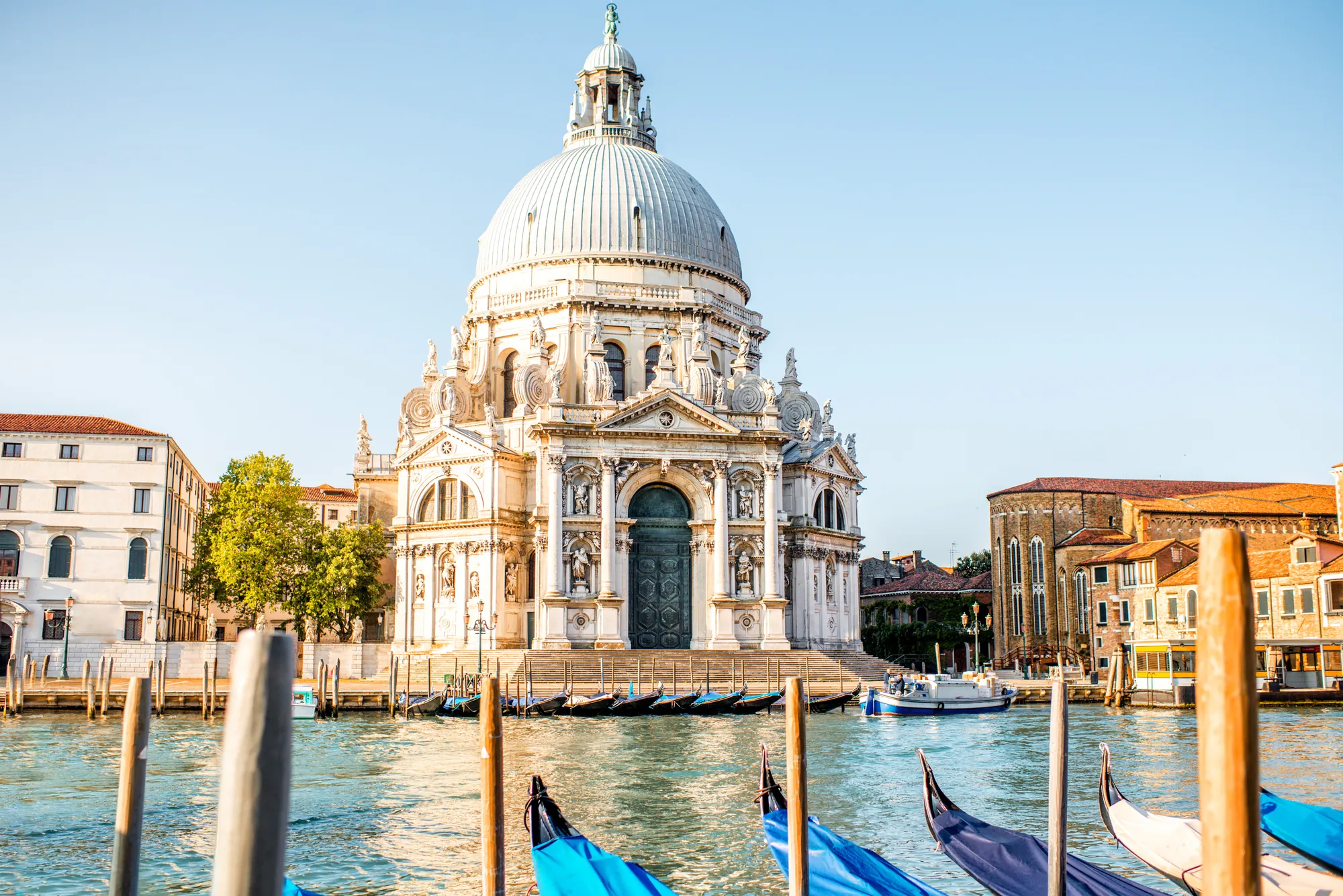 Nahaufnahme der Santa Maria della Salute in Venedig bei sonnigem Wetter, die im strahlenden Licht majestätisch am Ufer des Canal Grande steht. Im Vordergrund liegen Boote ruhig am Wasser, während die markante Kuppel der berühmten Kirche imposant über der Umgebung thront. Die Sonne spiegelt sich sanft auf dem Kanal, was der Szene einen besonderen Glanz verleiht.