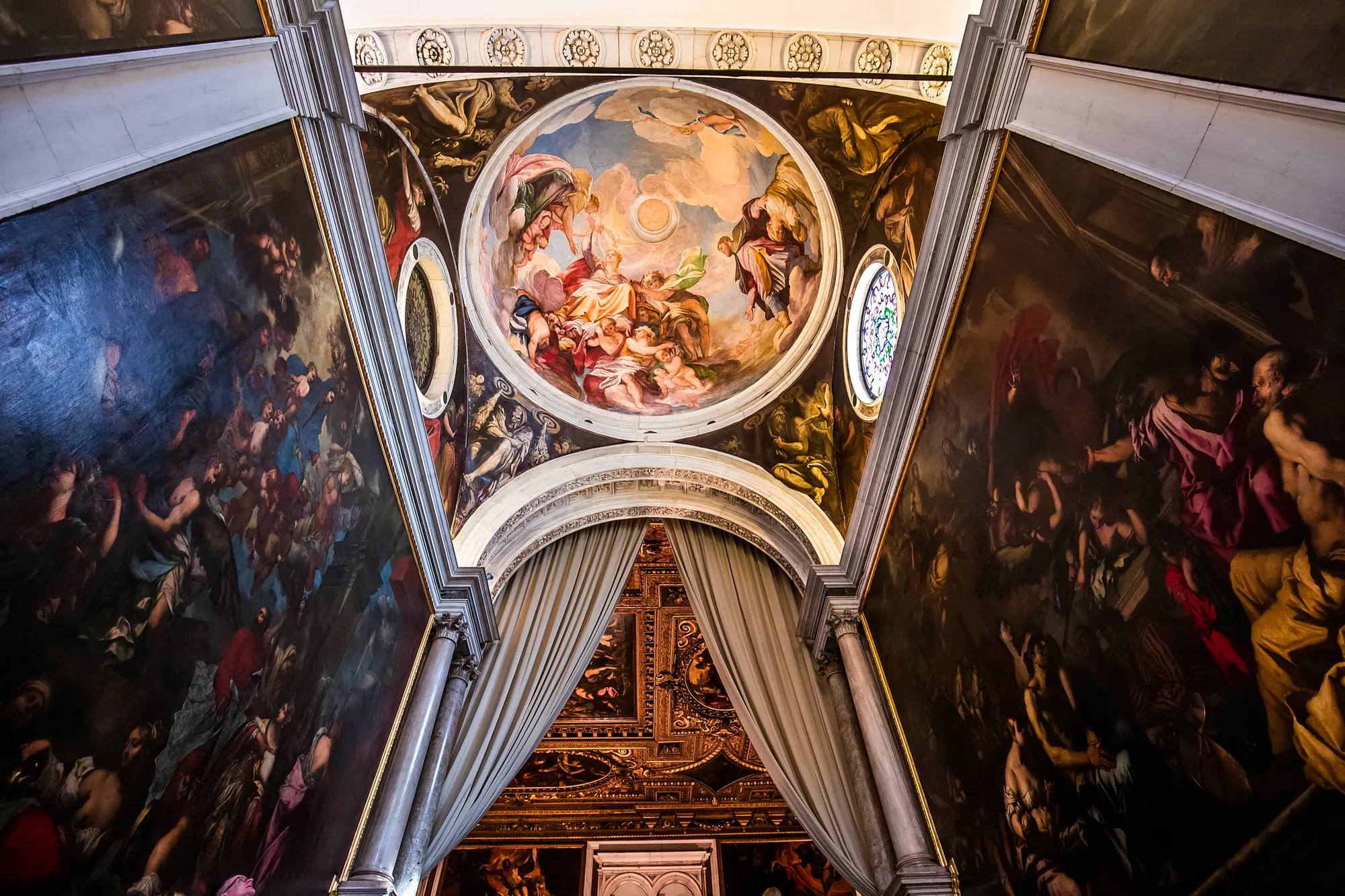 Innenansicht der Kuppel des Scuola Grande di San Rocco, reich verziert mit prachtvollen Fresken und kunstvollen Details. Das sanfte Licht fällt durch die Fenster und hebt die lebendigen Farben der Malereien hervor, die die heiligen Szenen und Symbolik des Ortes darstellen. Die kunstvolle Gestaltung der Kuppel schafft eine beeindruckende Atmosphäre, die die Besucher in die spirituelle und kulturelle Bedeutung dieses historischen Gebäudes eintauchen lässt.