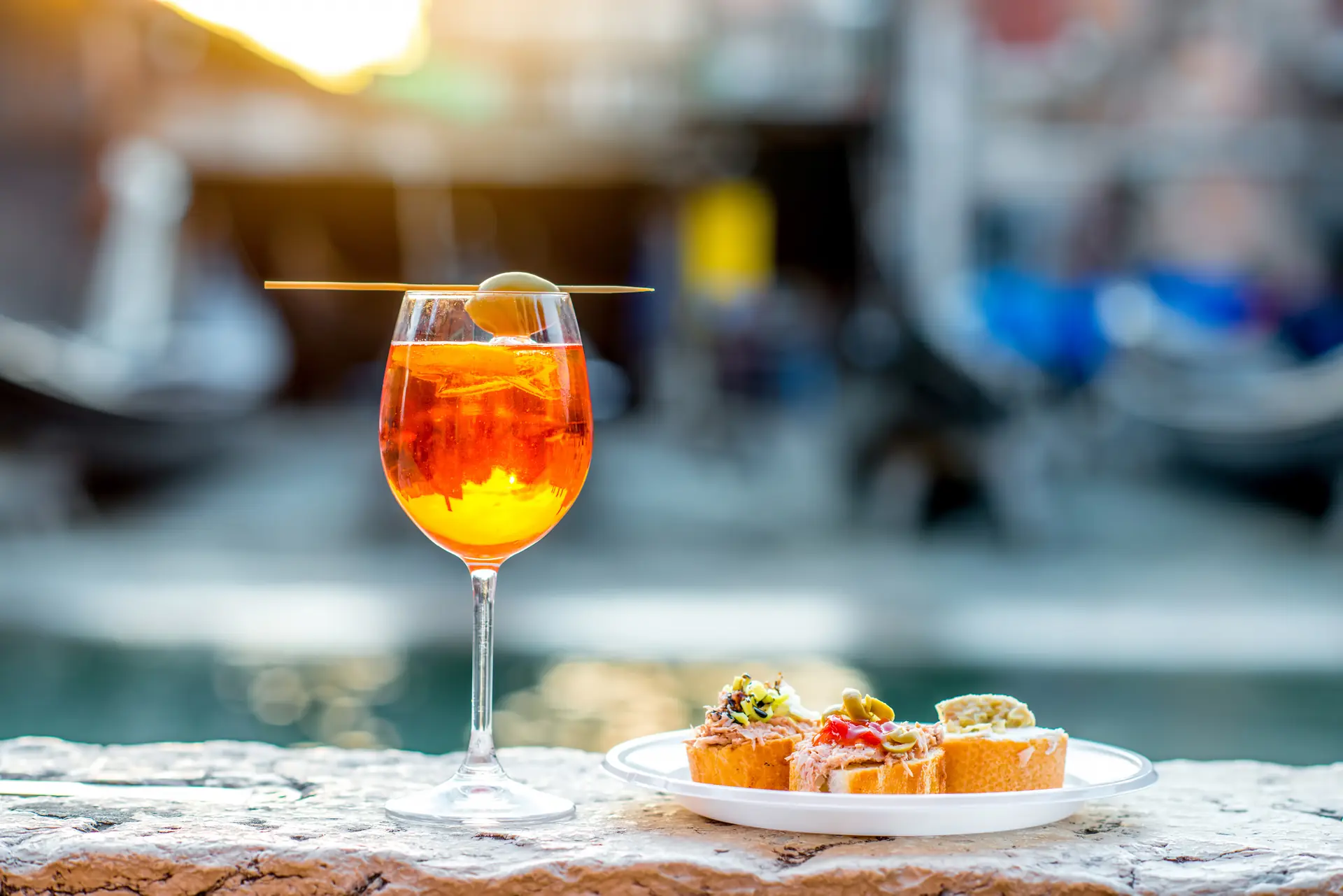 Nahaufnahme von Cicchetti und einem Aperol Spritz am Kanal in Venedig. Die appetitlichen Snacks sind ansprechend angerichtet, während der unscharfe Hintergrund die malerische Kulisse der Stadt andeutet, was die entspannte Atmosphäre eines italienischen Aperitifs unterstreicht.