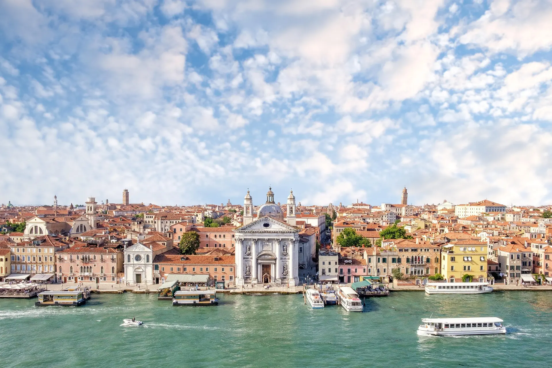 Im Vordergrund fließt der Canale della Giudecca, gesäumt von Wassertaxis und Wasserbussen in Venedig. Die Hafenpromenade ist belebt mit vielen Spaziergängern, die den sonnigen Tag genießen. Direkt an der Promenade befindet sich die Kirche Santa Maria del Rosario, umgeben von farbenfrohen Häusern, die das charmante Stadtbild unterstreichen.