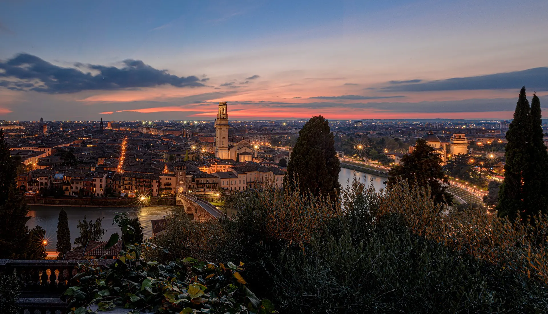 Panorama über Verona in leicht rosalicher Abendstimmung, das die Stadt mit ihrem Lichterglanz und den historischen Gebäuden sanft erleuchtet.