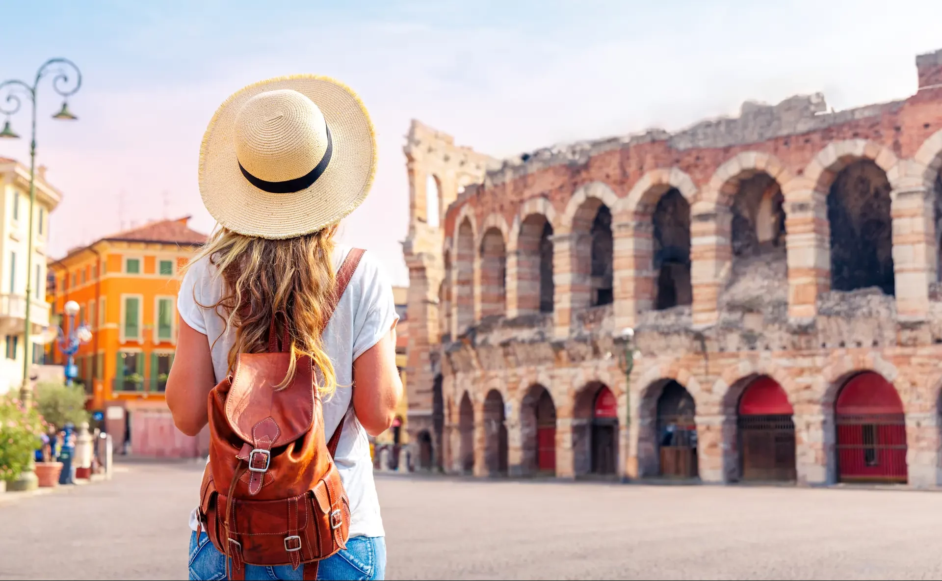 Eine Frau blickt auf die beeindruckende Arena di Verona bei mildem Wetter. Die majestätische römische Amphitheaterstruktur ist im Hintergrund sichtbar, umgeben von der historischen Architektur der Stadt, während der Himmel klar und einladend ist.