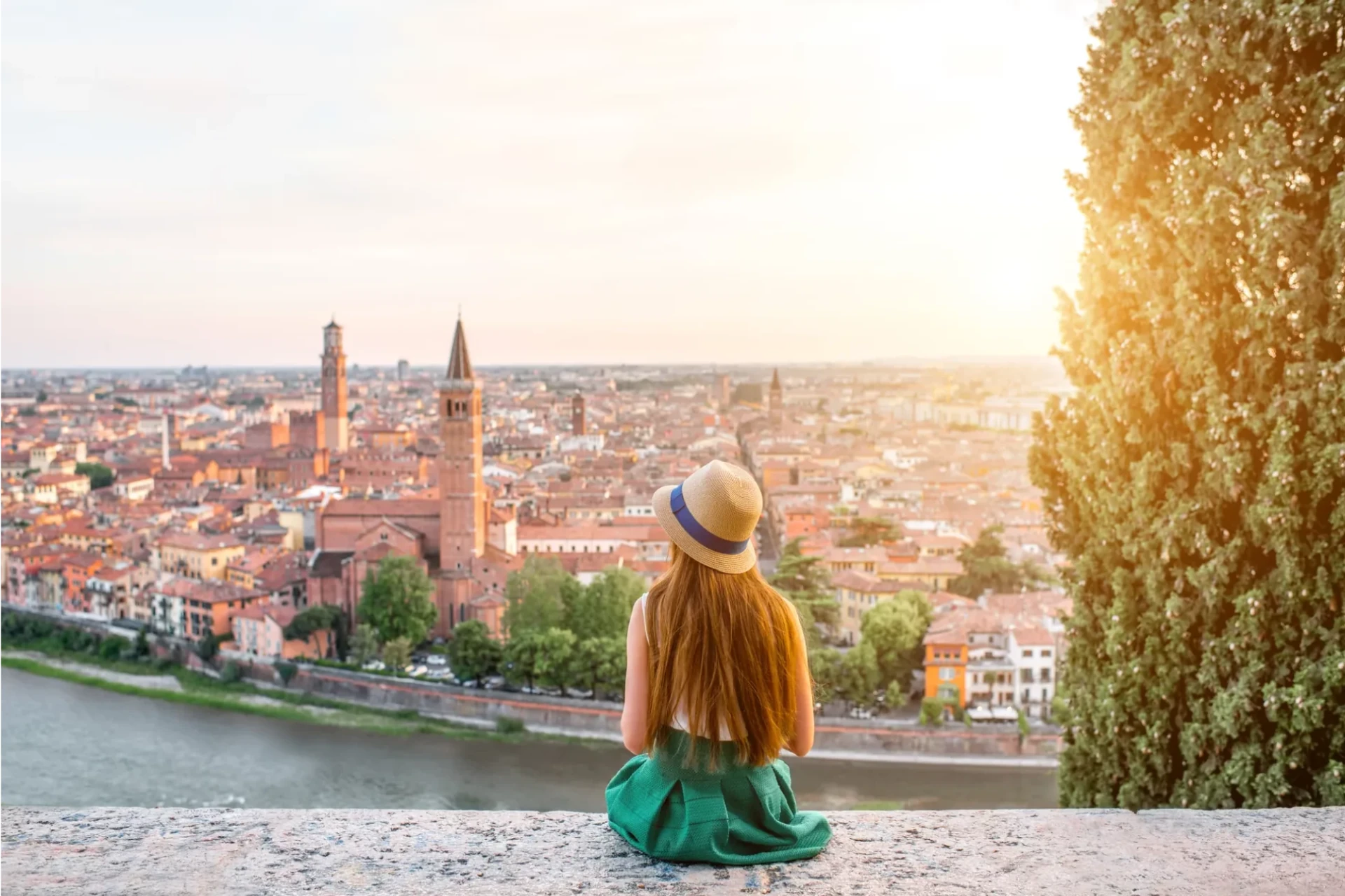 Eine Frau sitzt auf der Mauer und blickt über den Etsch auf die Stadt Verona, während ein sanfter Sonnenuntergang die Szene in warmes Licht taucht. Die malerische Skyline der Stadt ist im Hintergrund zu sehen, umgeben von der natürlichen Schönheit der Umgebung.