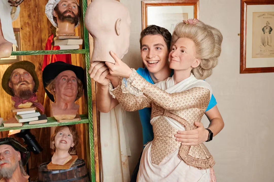 Wachsfigurenkabinett Madame Tussauds Berlin - Marie Tussaud betrachtet stolz eine modellierte Büste. Im Hintergrund sind ein Regal mit weiteren Büsten zu sehen und ein junger Mann hält den linken Arm um Madame Tussauds Taille und schaut ebenfalls auf die neue Büste.