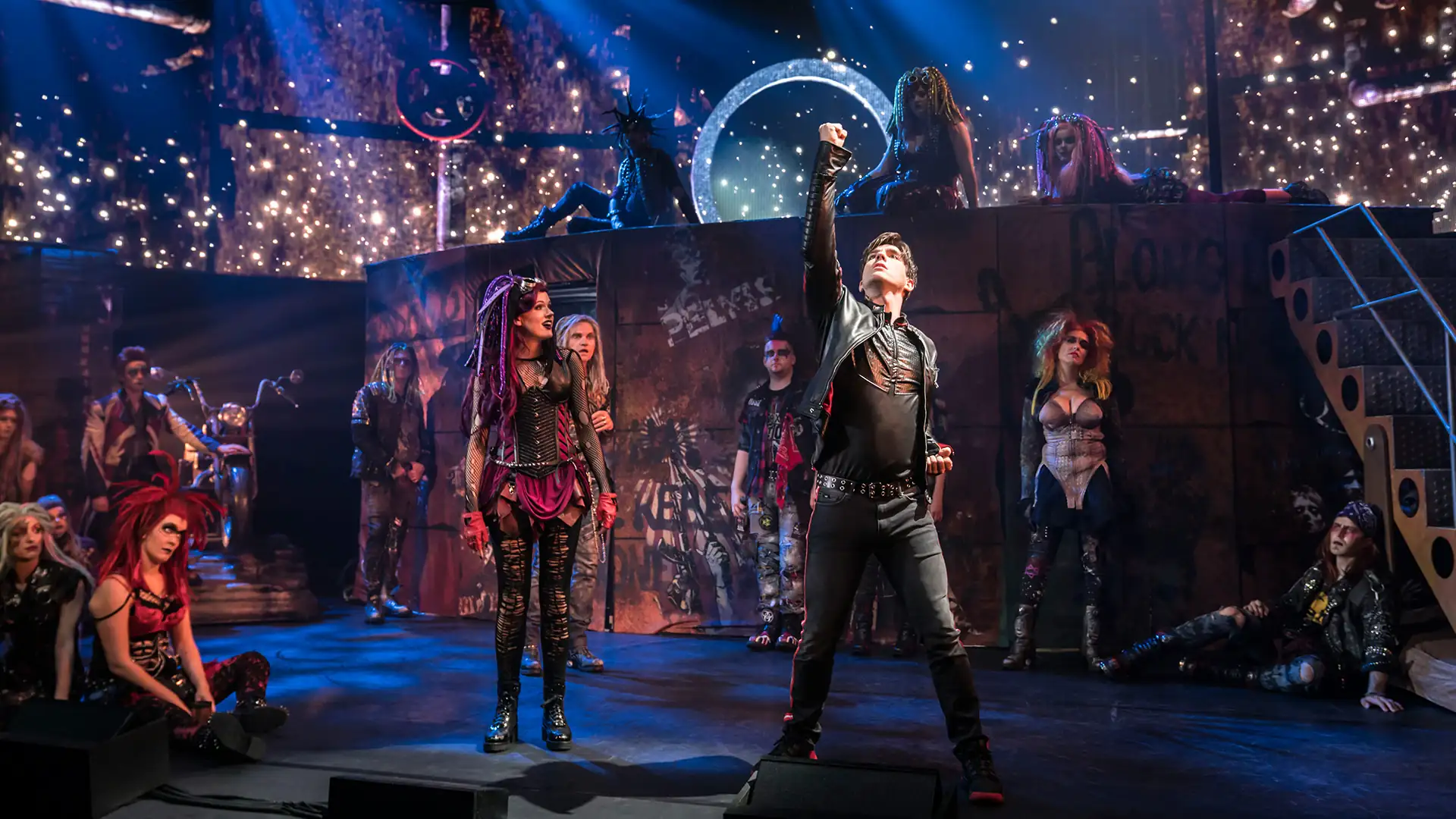We will Rock You – Das Musical (Bild im Kachelformat), Szenenbild: Darsteller in schwarzer Kleidung reckt die Faust in die Höhe im dystopischen Setting. (UK Cast)