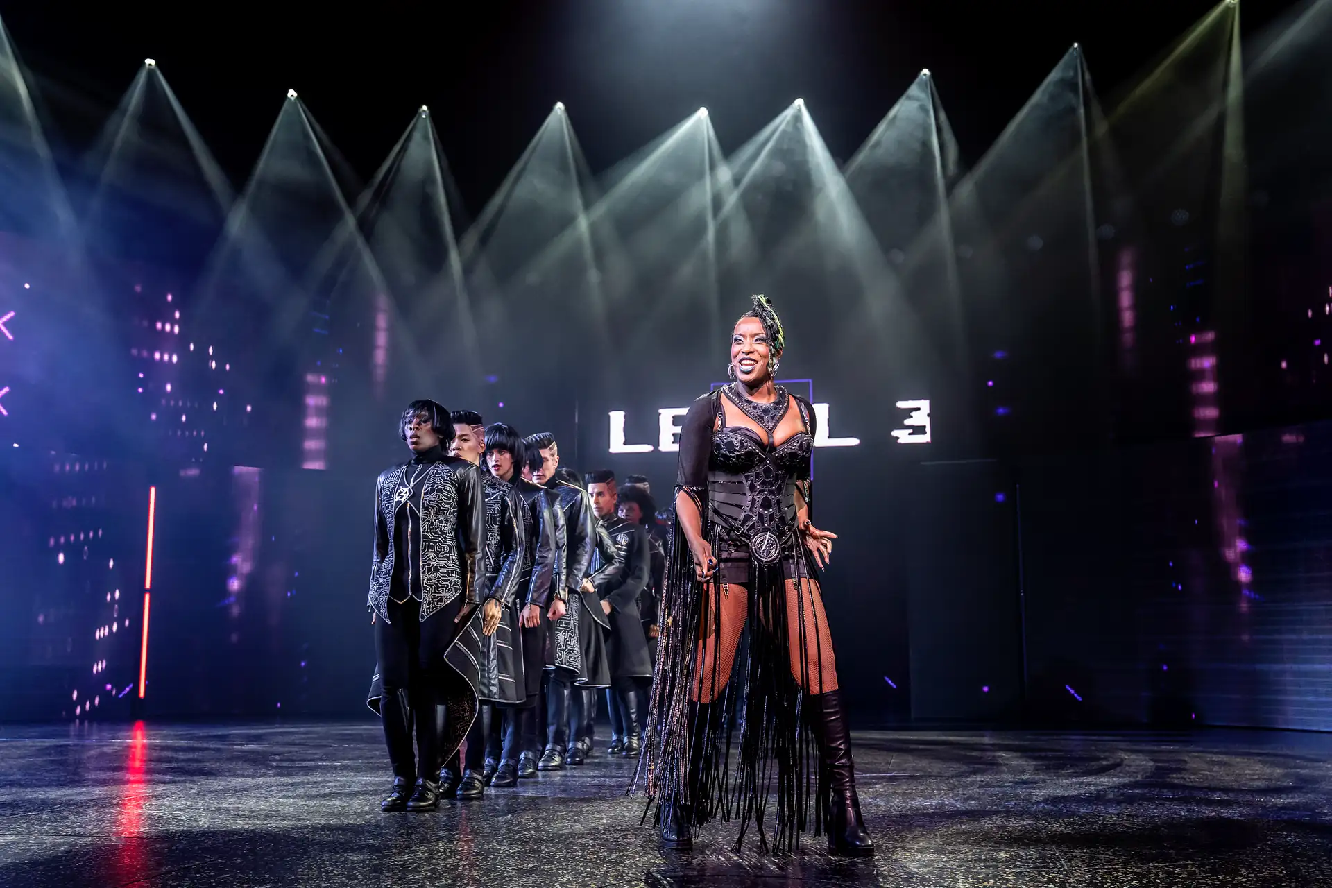 We will Rock You – Das Musical in Stuttgart, Szene: Aisata Blackman als Killer Queen in schwarzem, transparentem Kleid mit Fransen steht vorn auf Bühne, dahinter mehrere Personen in dunkler Kleidung, Bühnenbeleuchtung und Schriftzug 'LEVEL 3'.