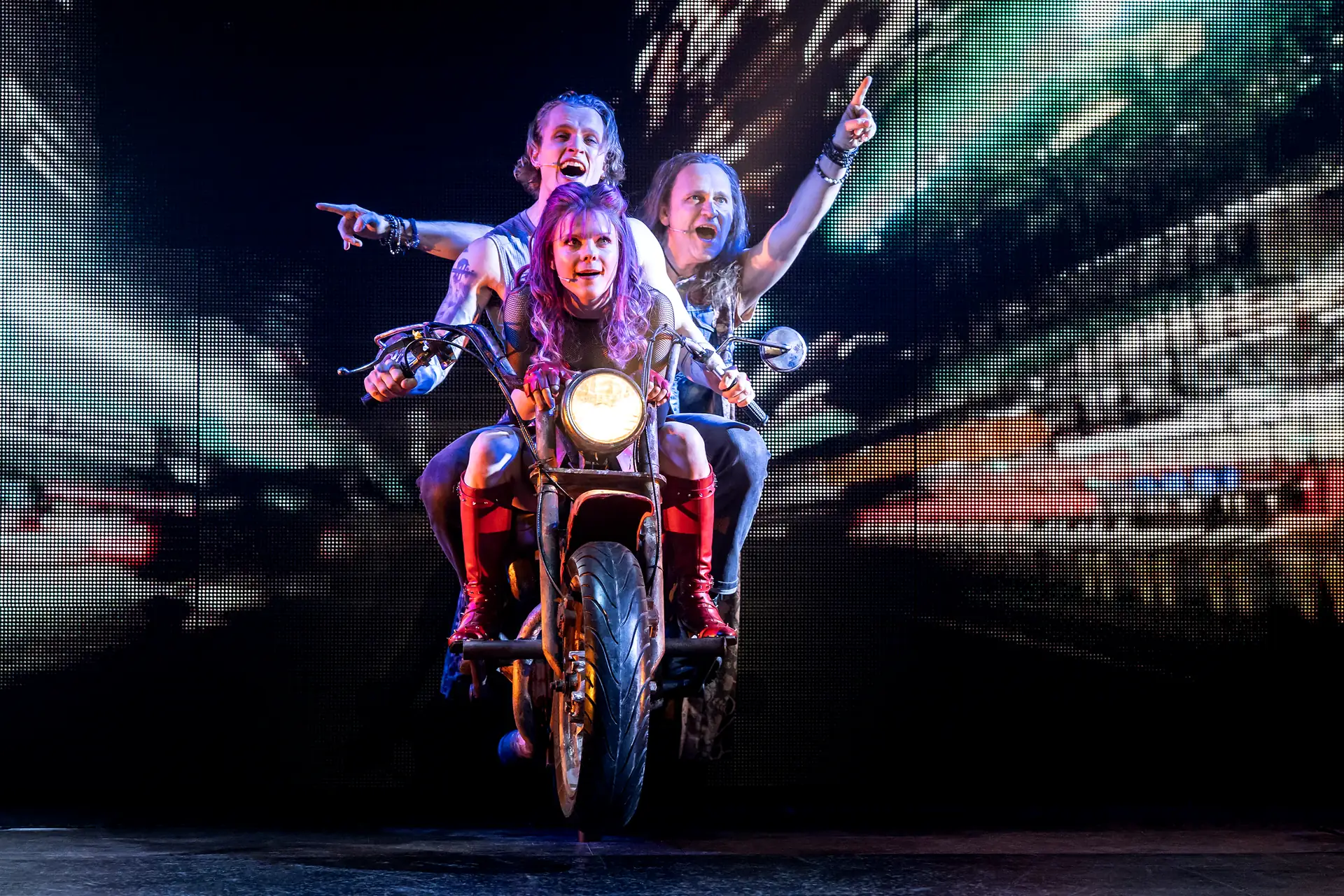 We will Rock You – Das Musical in Stuttgart, Szene: Drei Personen auf einem Motorrad, die vordere Person hält den Lenker, die hinteren heben jeweils einen Arm und zeigen mit dem Finger nach oben.