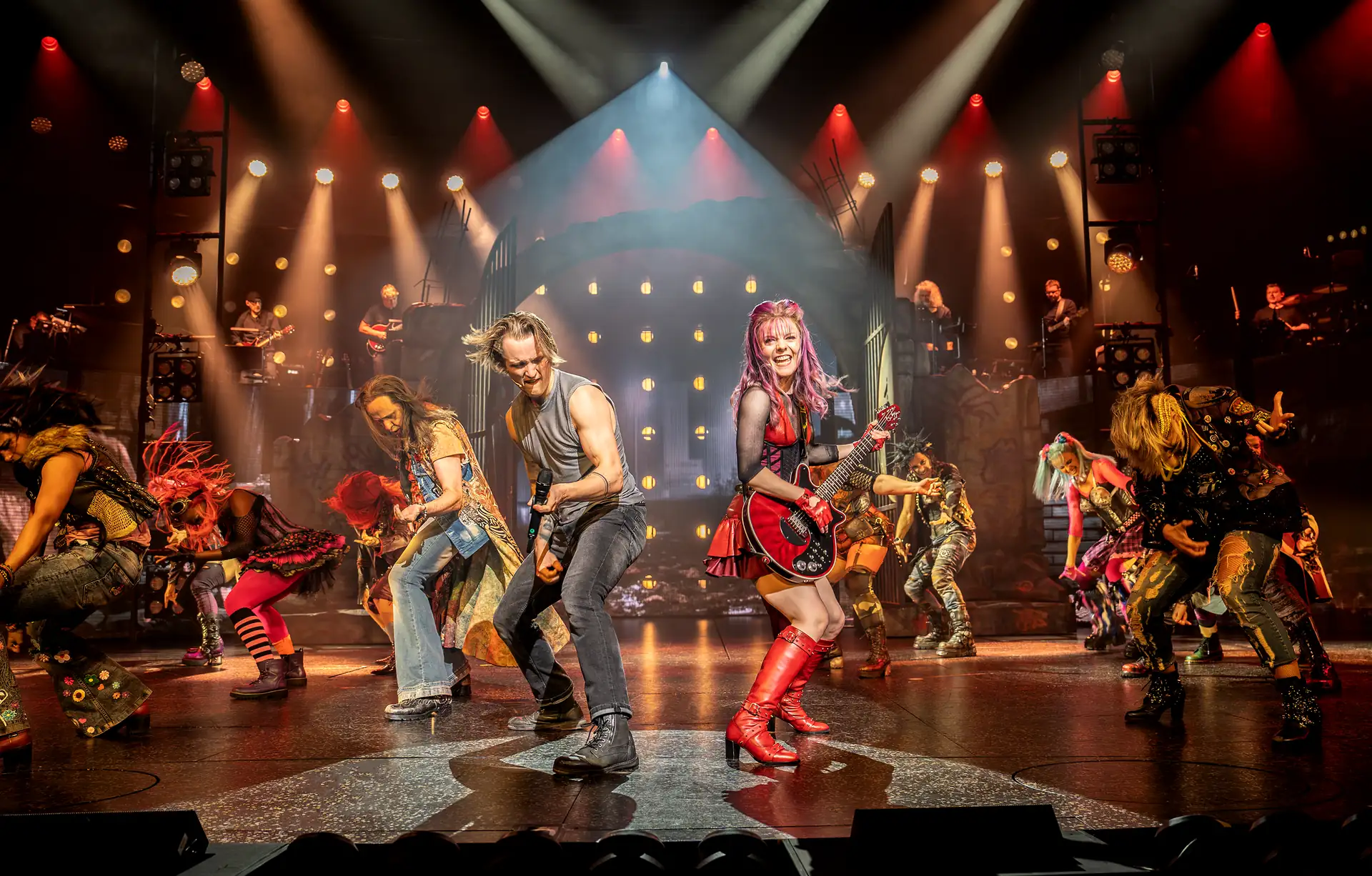 We will Rock You – Das Musical in Stuttgart, Szene: Bühnenbild mit mehreren Musikern und Tänzern in bunten Kostümen, darunter eine Frau mit rotem Kleid und roten Stiefeln, beleuchtet von Scheinwerfern.