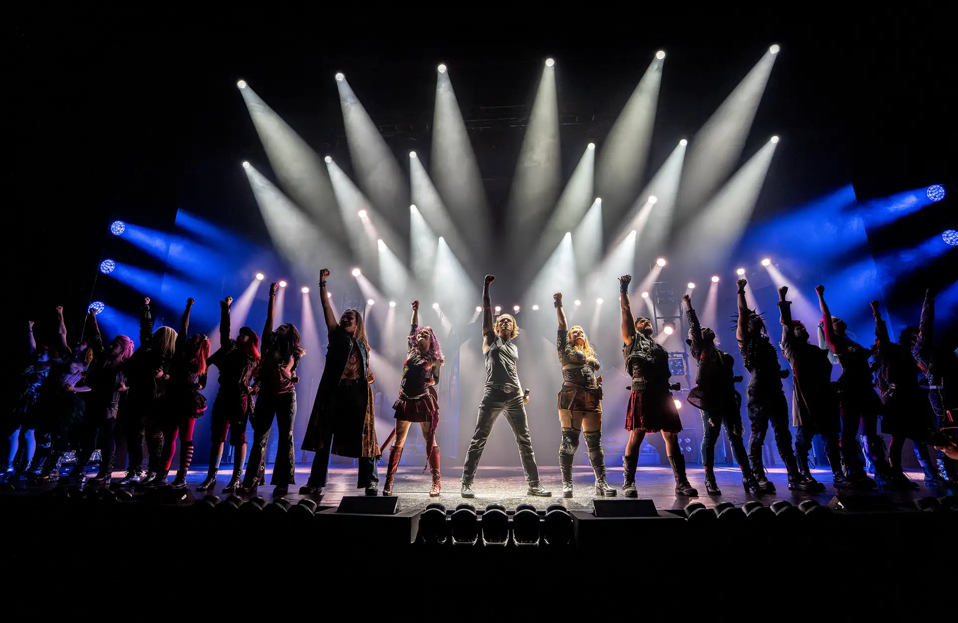 We will Rock You – Das Musical in Stuttgart, Szene: Menschen auf Bühne mit erhobenen Fäusten, beleuchtet von weißen und blauen Scheinwerfern.