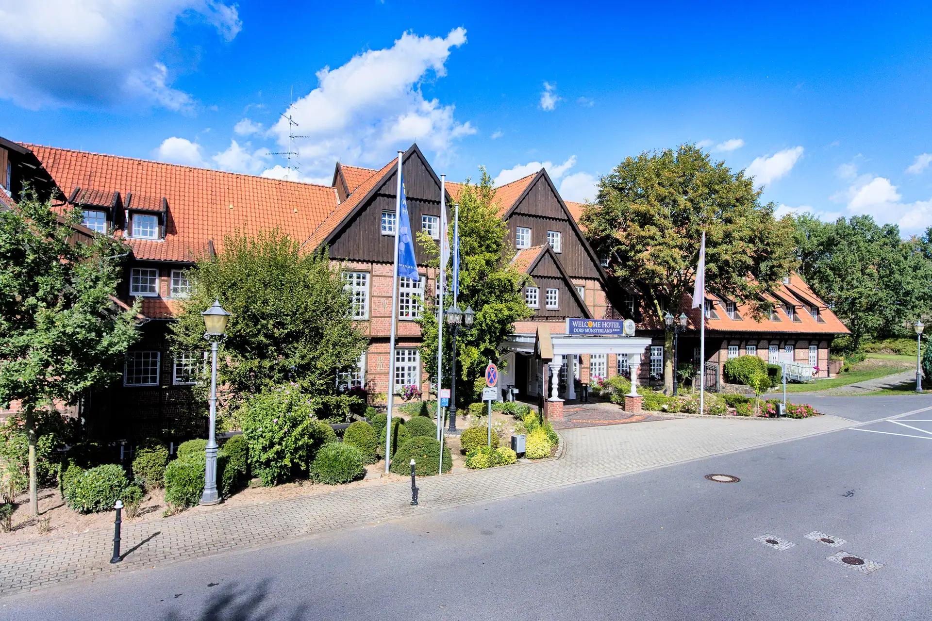 Außenansicht des Welcome Hotel Legden mit Fachwerkgebäuden, roten Ziegeldächern, Fahnenmasten und begrünten Eingangsbereich an einer ruhigen Straße