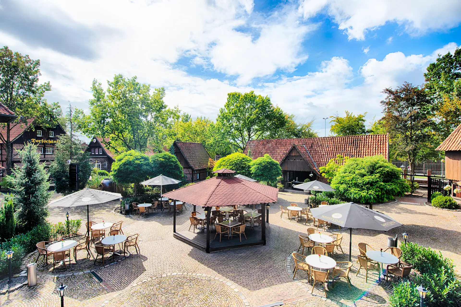 Biergarten des Welcome Hotel Legden mit mehreren runden Tischen, Stühlen und Sonnenschirmen auf gepflastertem Platz, umgeben von Fachwerkhäusern und Bäumen