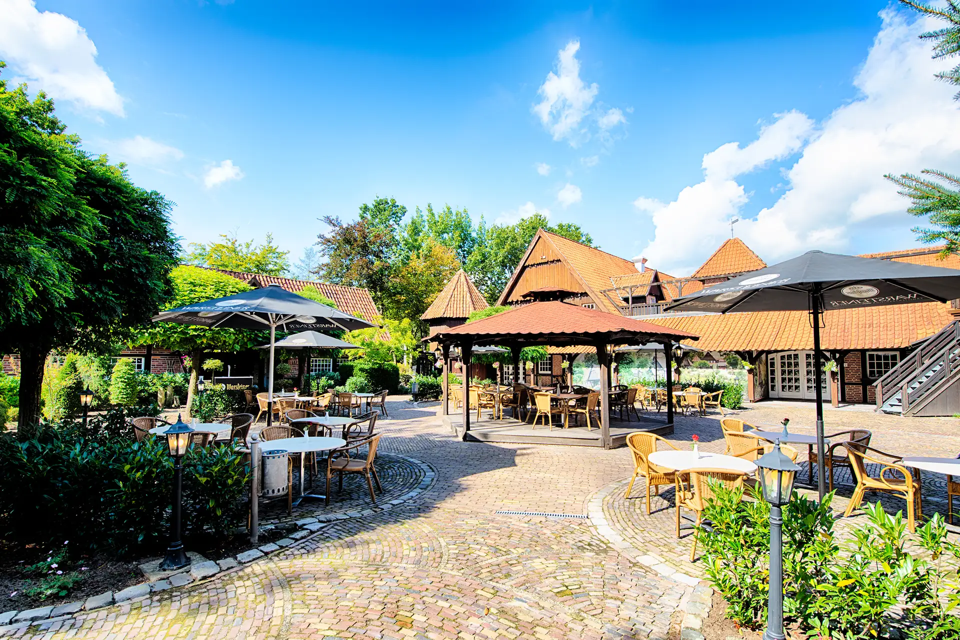 Biergarten mit gepflastertem Boden, mehreren Tischen und Stühlen unter großen Sonnenschirmen sowie einem hölzernen Pavillon vor dem Welcome Hotel Legden