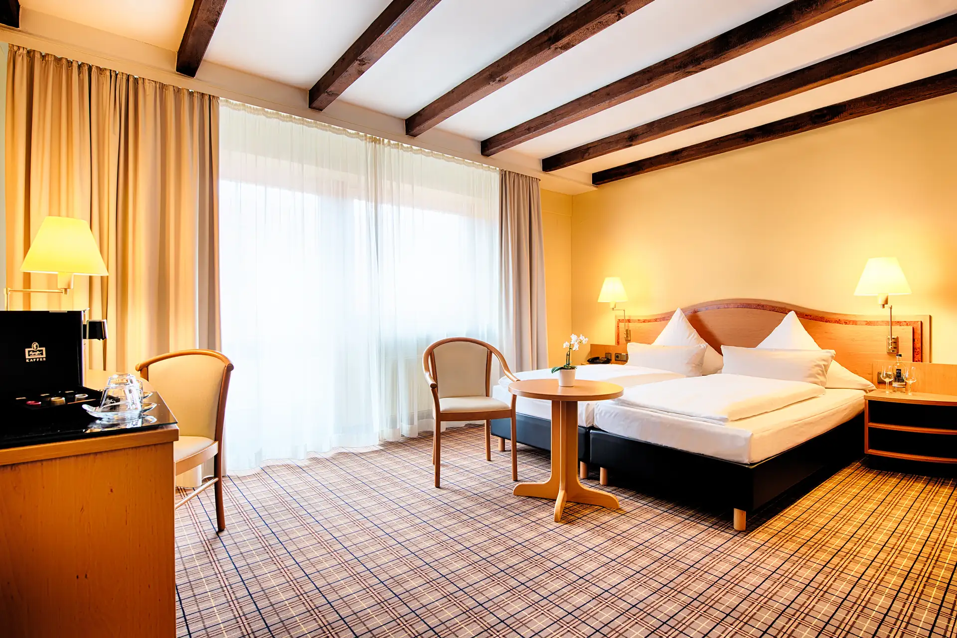 Hotelzimmer mit Doppelbett, zwei Sesseln, kleinem runden Tisch, Schreibtisch mit Stuhl und Stehlampe, Teppichboden und sichtbaren Holzbalken an der Decke