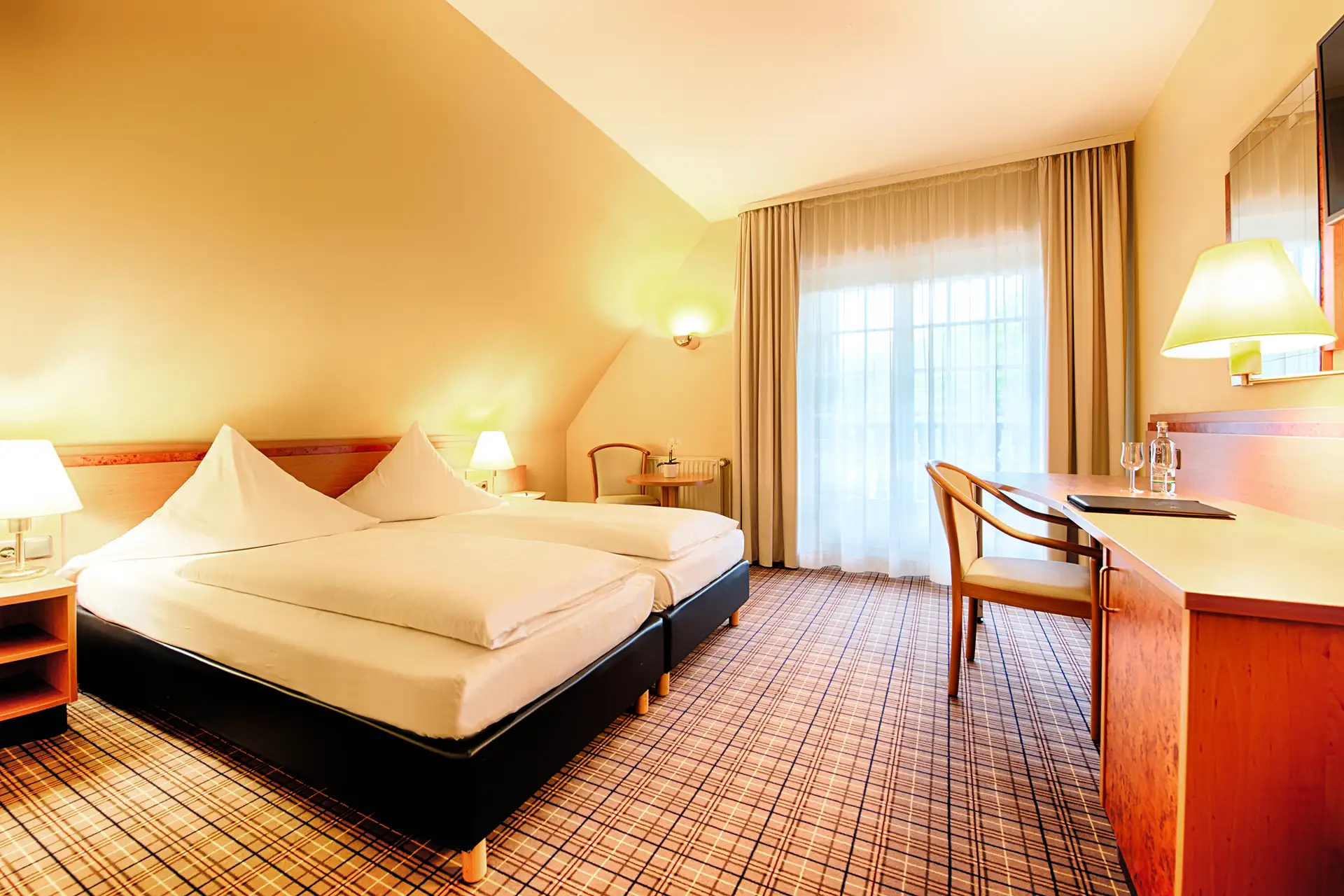 Hotelzimmer mit zwei Einzelbetten, Nachttisch mit Lampe, Schreibtisch mit Stuhl und großer Fensterfront mit Vorhängen