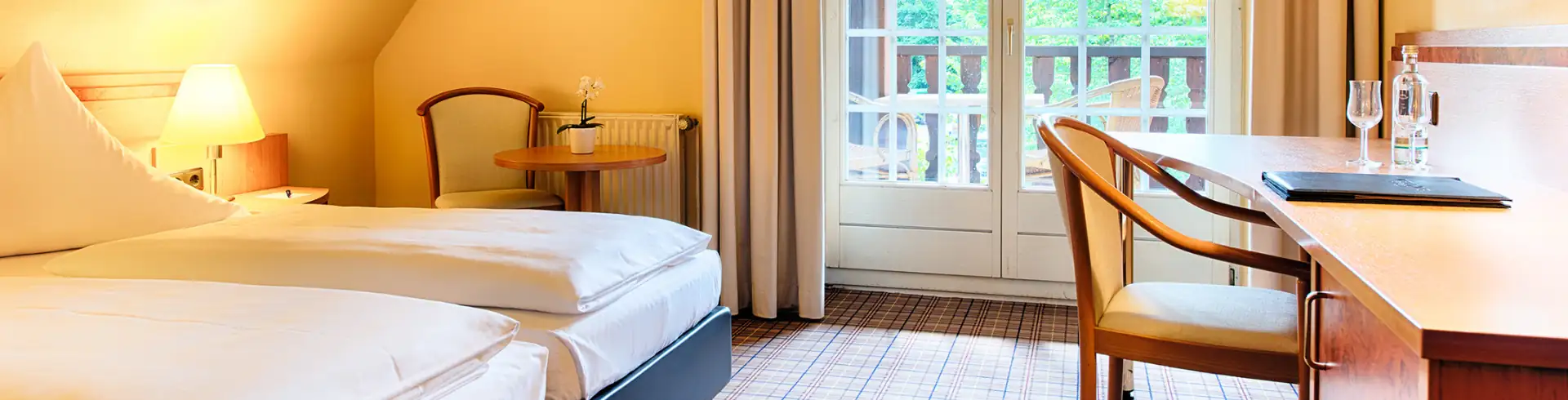 Hotelzimmer mit zwei Einzelbetten, Nachttisch mit Lampe, Stuhl, Schreibtisch mit Stuhl und Balkon mit Geländer im Hintergrund (im Bannerformat)