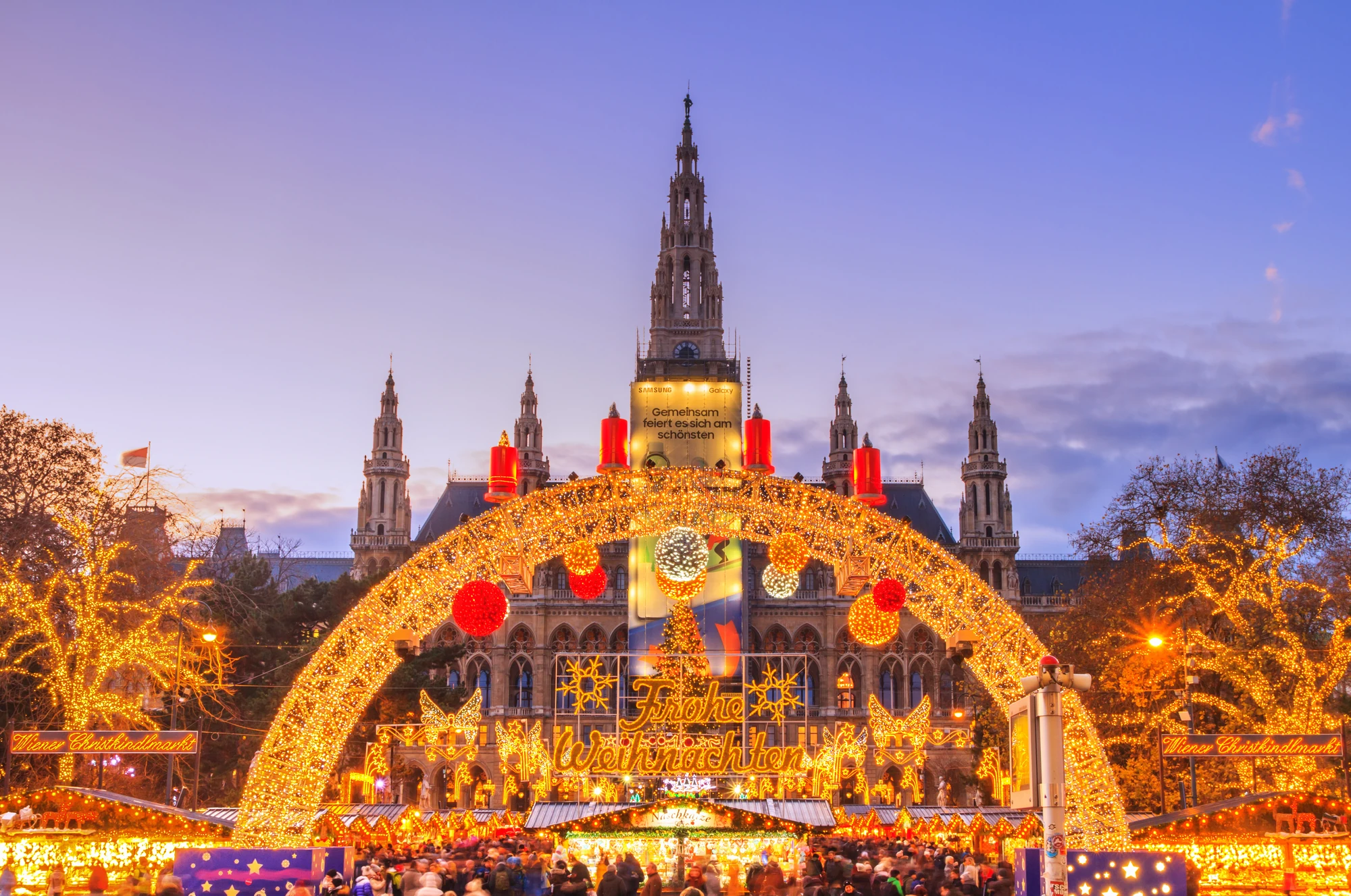 Beleuchteter Weihnachtsmarkt mit großem Lichterbogen und Rathaus im Hintergrund bei Abenddämmerung in Wien