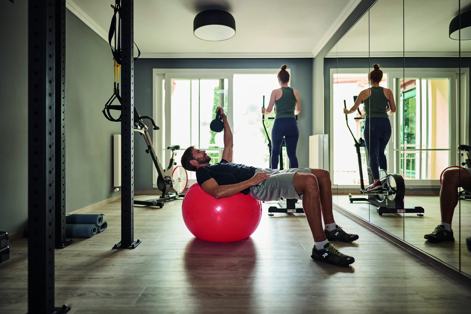 Das Bild zeigt einen modernen Fitnessraum im aja Strandhotel Bansin, ausgestattet mit hochwertigem Trainingsequipment wie einem Gymnastikball, einem Crosstrainer und einer Kraftstation. Die lichtdurchflutete Atmosphäre mit großen Fenstern und Spiegelwänden bietet ideale Bedingungen für ein effektives und entspanntes Workout.